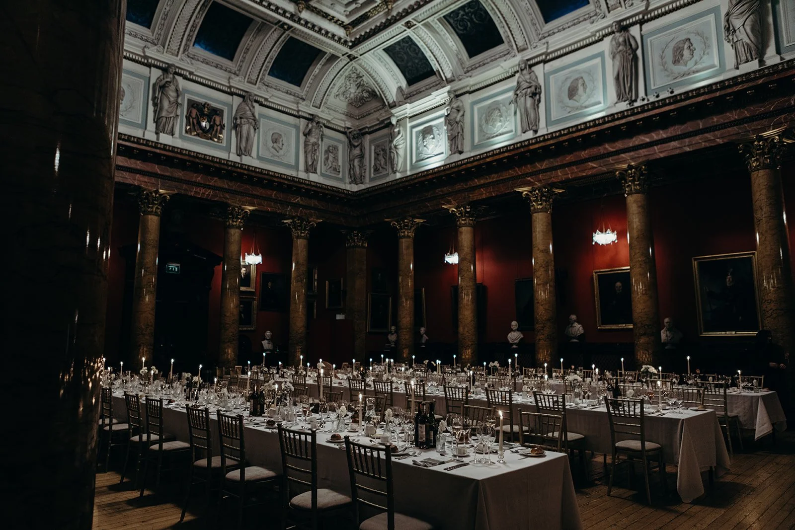 Elegant dining hall decorated for a formal event with long tables set with glassware, cutlery, and napkins, ornate gold and marble columns, red walls, portraits and bust sculptures, and chandeliers illuminating the space.