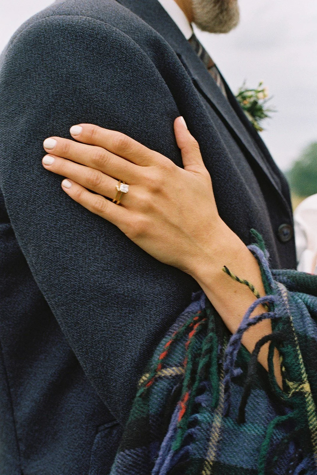 Close-up of a woman's hand with an engagement ring resting on a man's shoulder, who is wearing a dark jacket with a plaid shirt underneath.