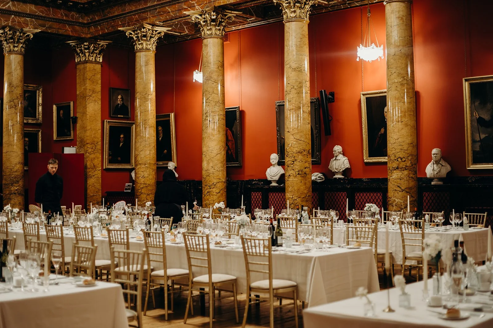 Elegant dining hall with white-covered tables, gold chairs, and tall marble columns; red wall with framed portraits and bust sculptures.