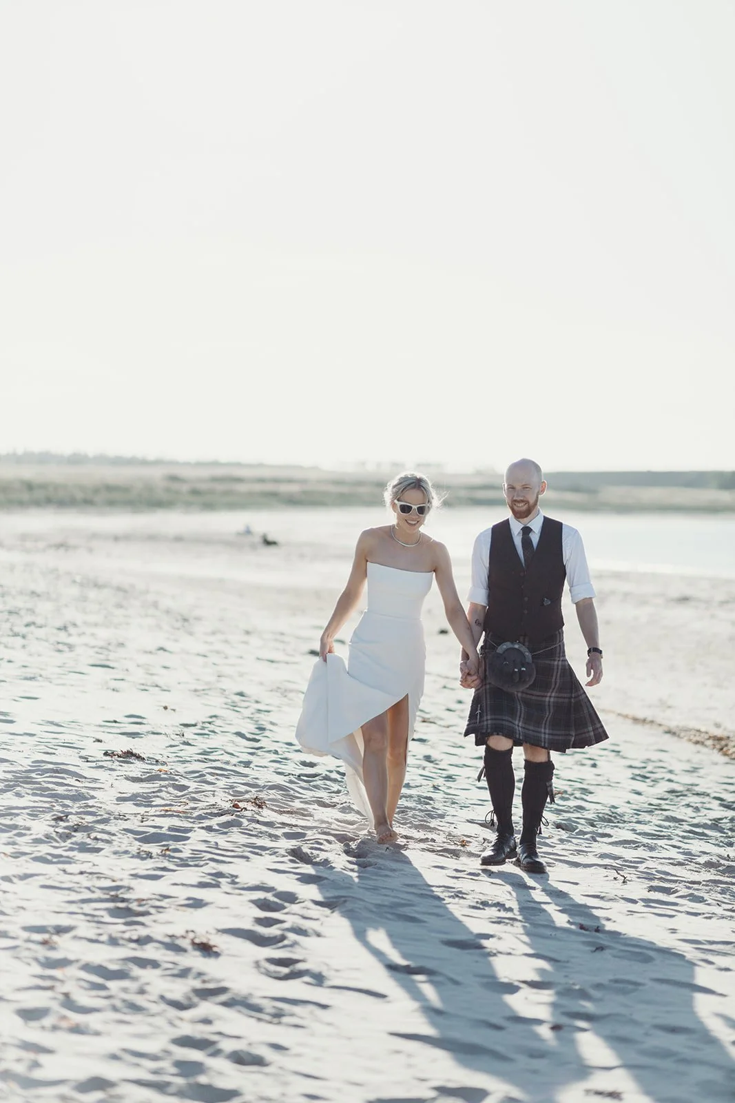 A couple walking hand in hand on the beach, dressed in wedding attire, with the woman in a white dress and the man in a kilt, both smiling.