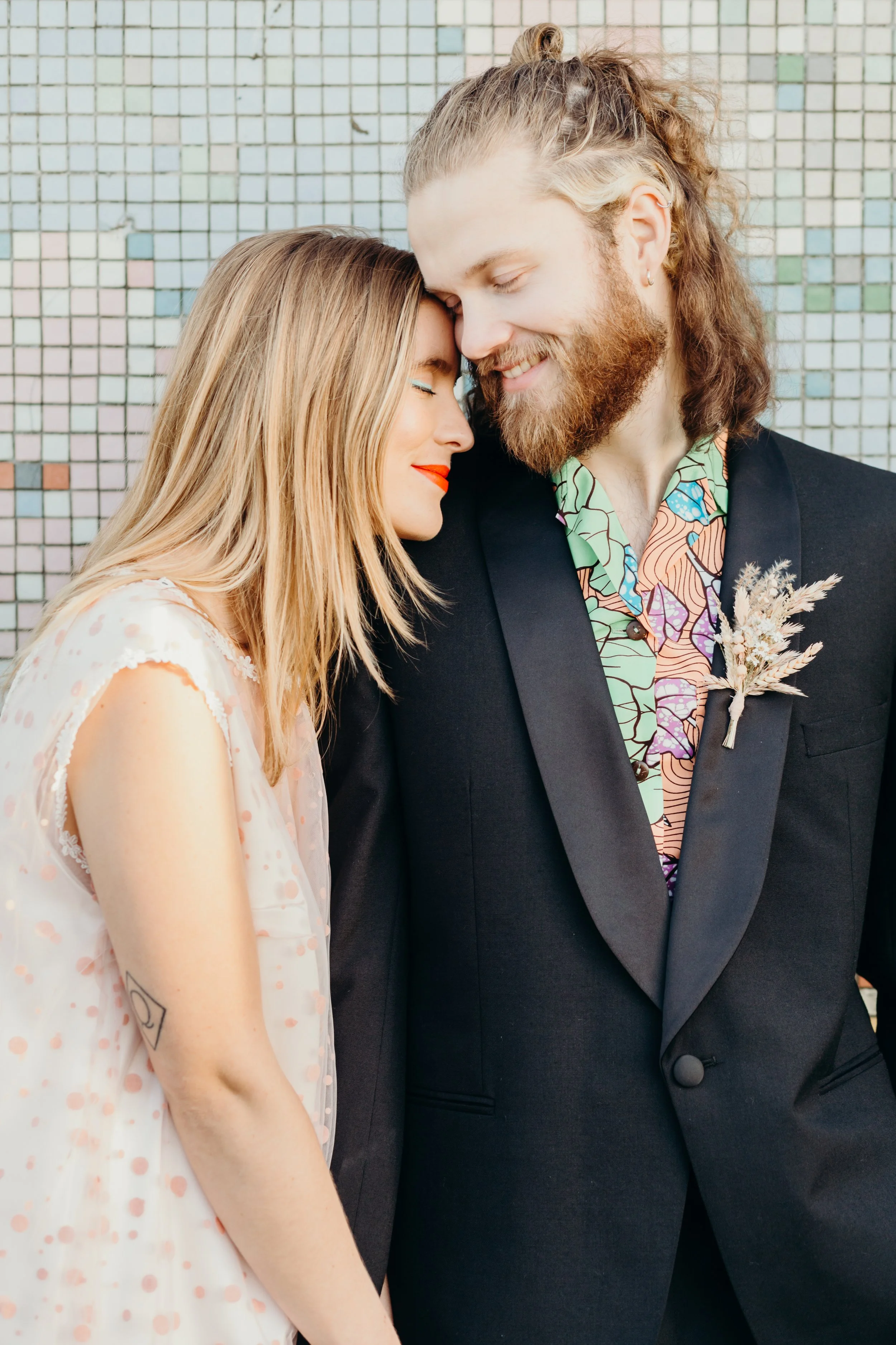 A man and woman with closed eyes and gentle smiles, standing close together, head touching, against a tiled wall.