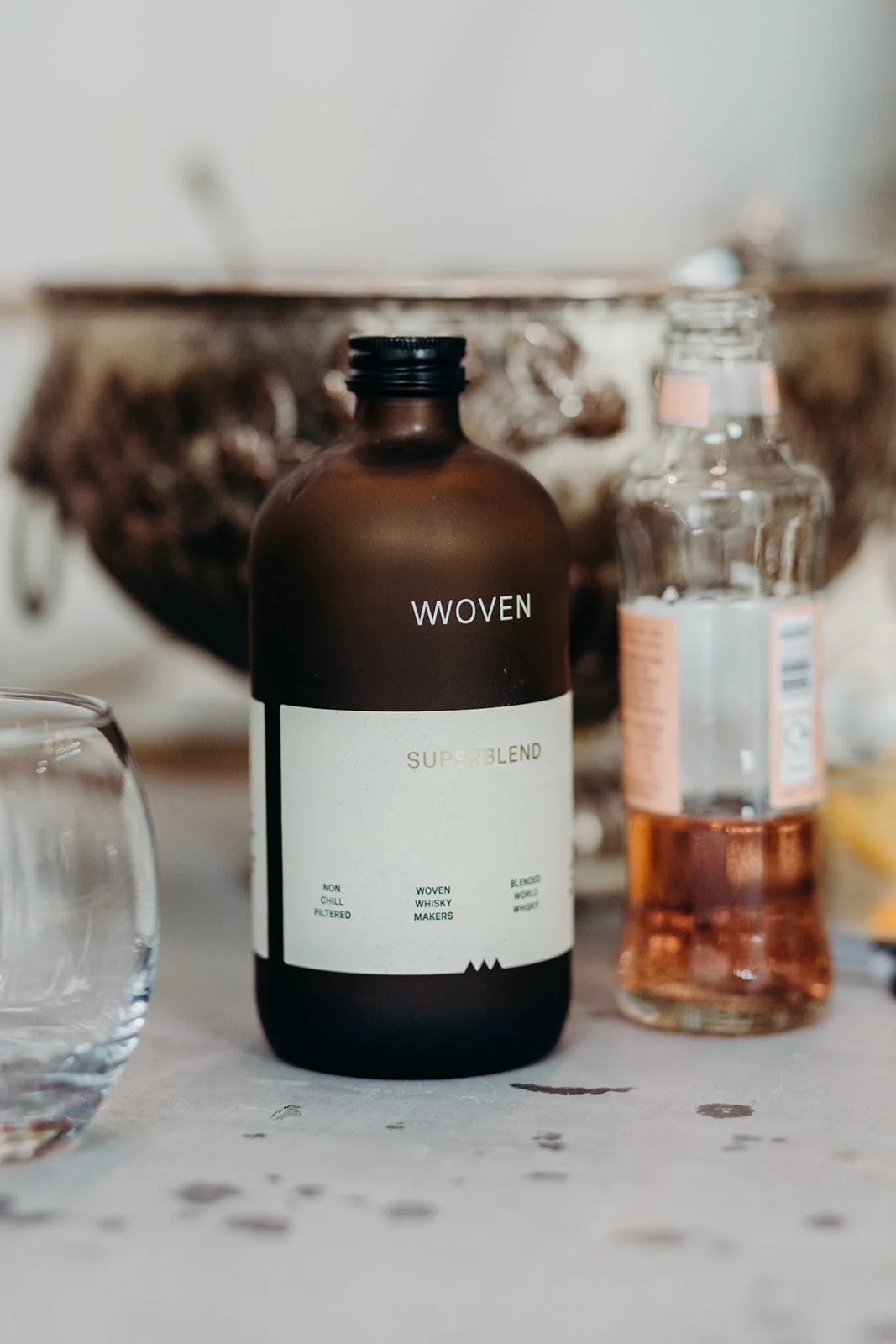 A brown bottle labeled 'WOVEN SUPERBLEND' next to a clear bottle with pink liquid and a lemon slice, surrounded by two glasses on a light surface with a rustic bowl in the background.