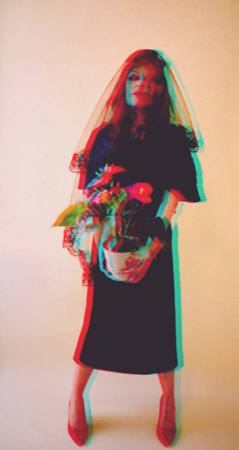 A woman in a black dress holding a potted plant with red and pink flowers, wearing a colorful mesh veil over her face.
