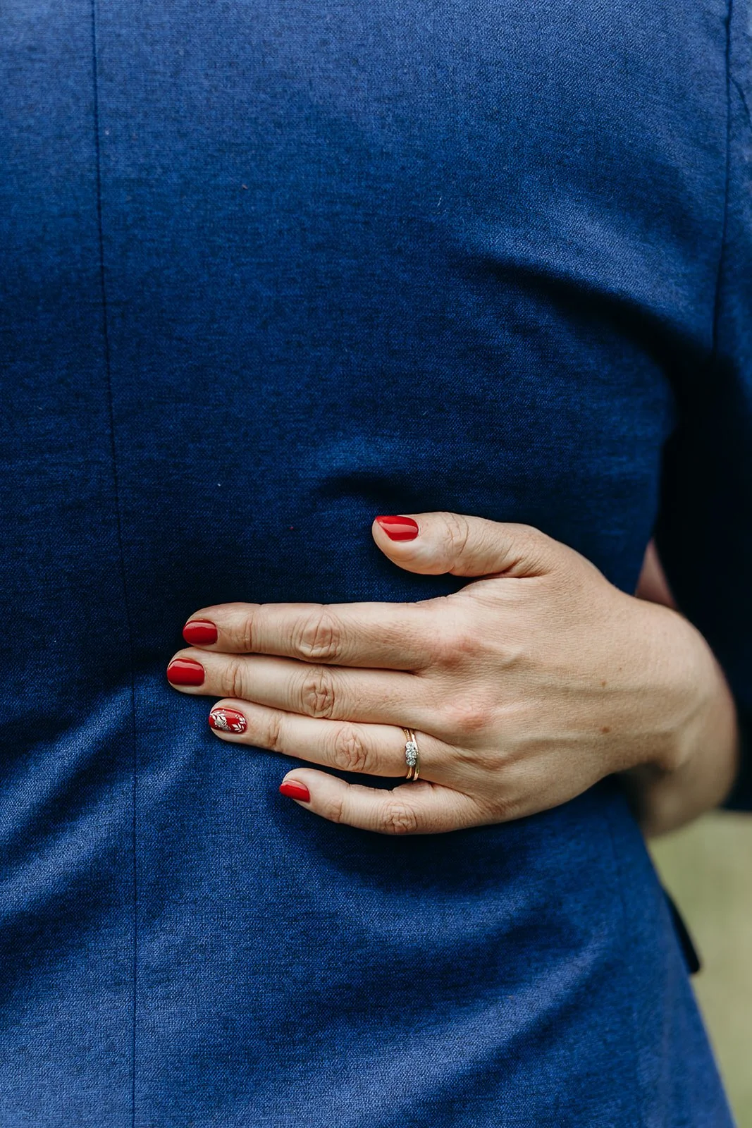 Close-up of a person's hand with red nails and a wedding ring, resting on their blue dress or suit.