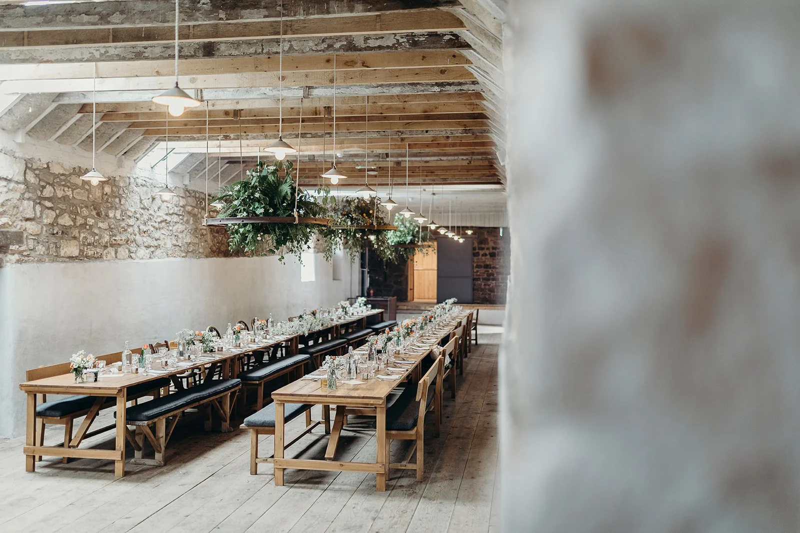 Long wooden banquet tables set with white flower arrangements, glassware, and place settings in a rustic barn with exposed wooden beams, stone walls, and soft lighting.