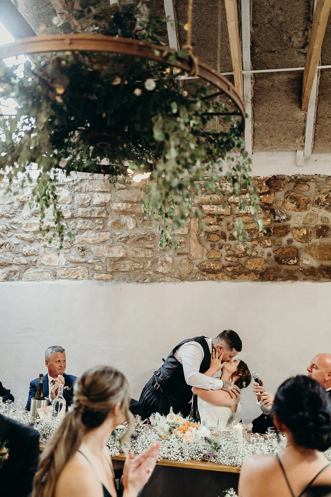 A wedding reception scene with a bride and groom sharing a kiss, surrounded by seated guests, flowers, and rustic stone wall in the background.