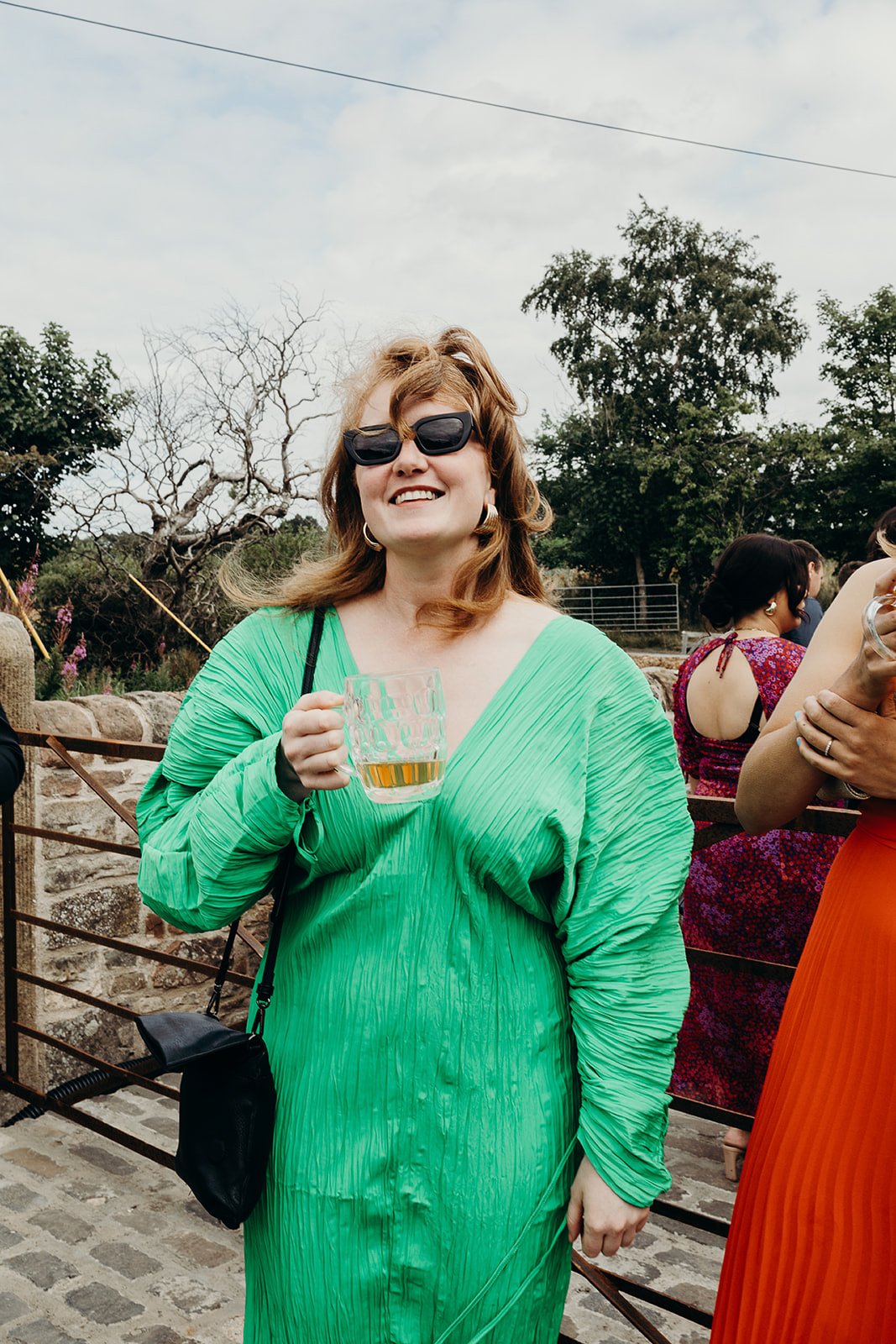 Woman in a bright green dress holding a glass of beer, smiling outdoors among other people, with trees and cloudy sky in the background.