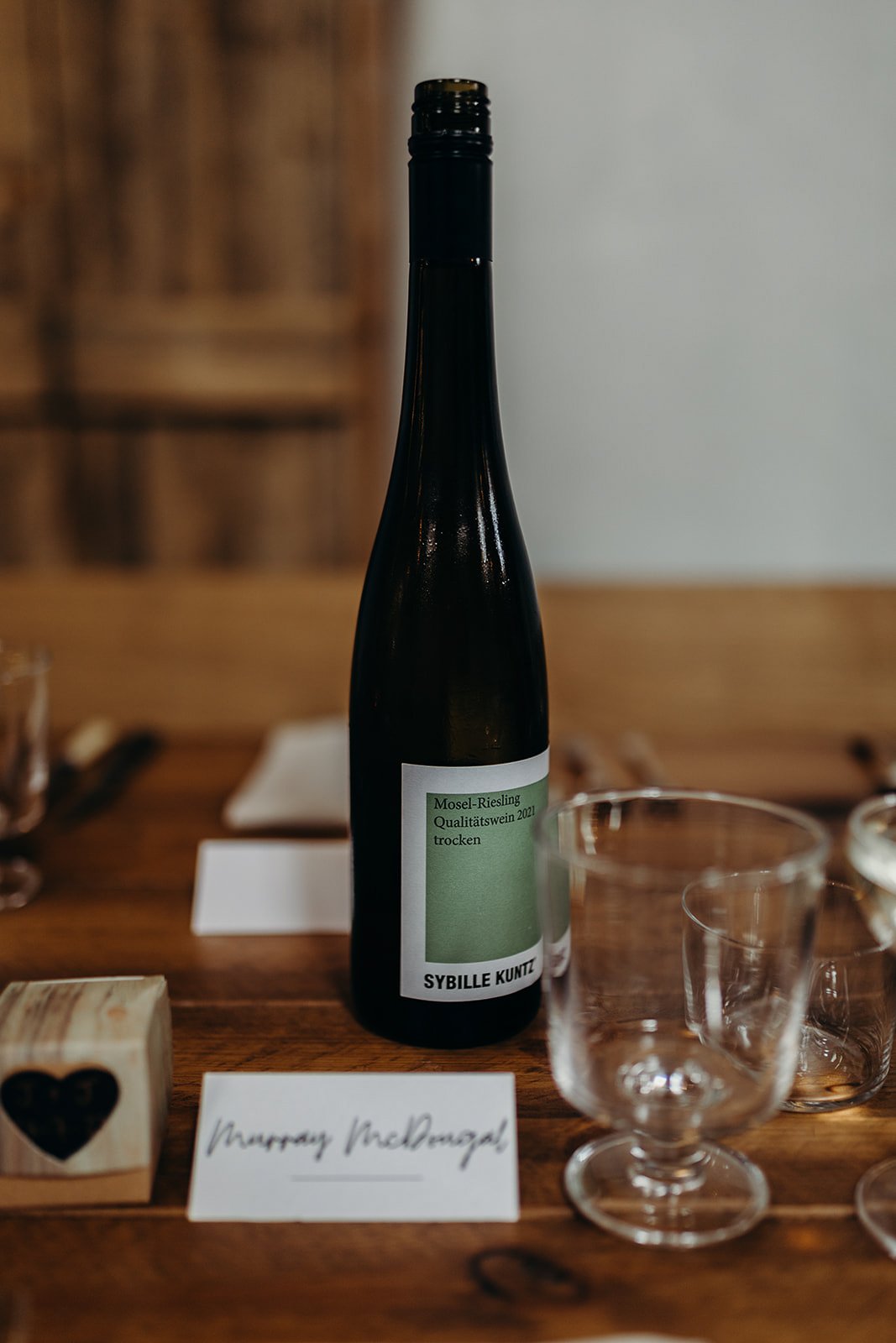 A bottle of Mosel Riesling Qualitätswein 2021 trocken wine on a wooden table, surrounded by empty glassware and a white card with handwritten text.