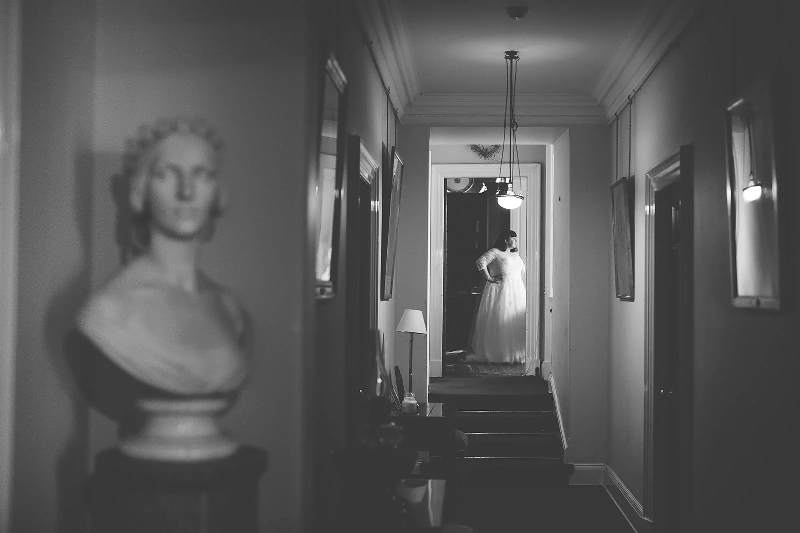 Black and white photo of a hallway with a woman in a wedding dress standing at the end, looking to the side, framed by a doorway. A blurred bust sculpture is in the foreground.
