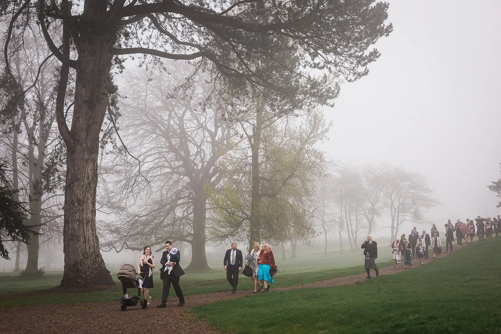 A group of people walking along a foggy park path surrounded by large trees.