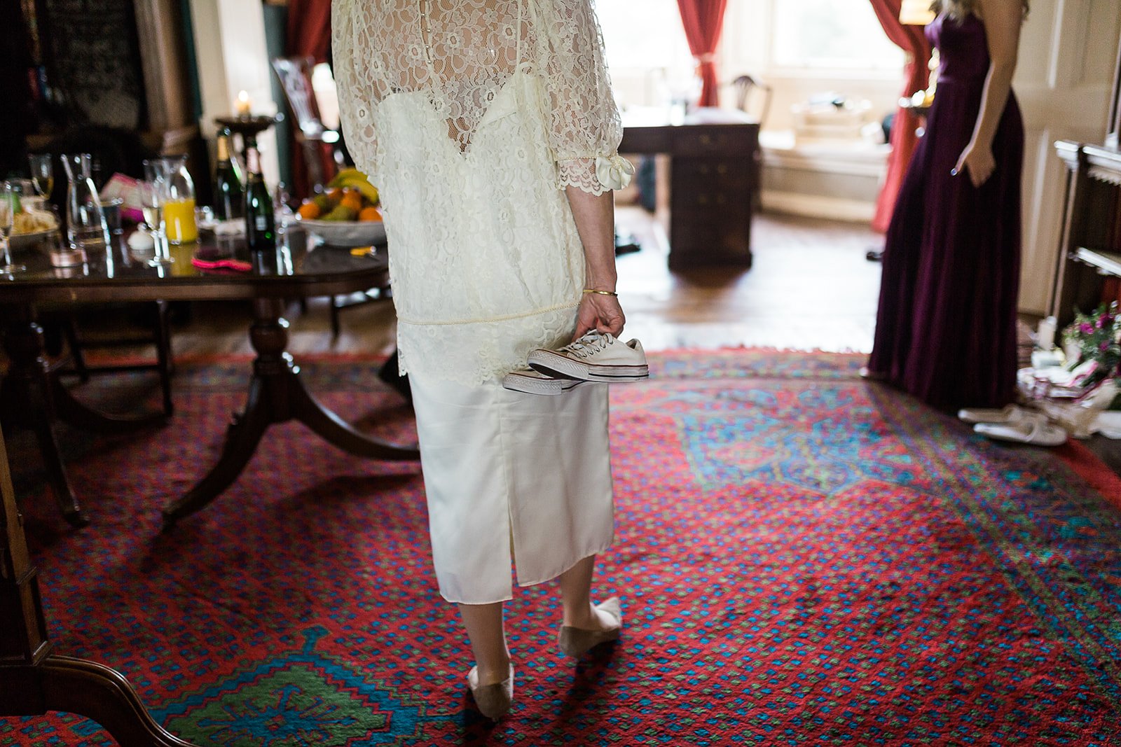 Woman in a lace dress holding a pair of white sneakers in her hand, standing on a multicolored patterned rug in a room with a table of food and drinks, and another woman in a dark purple dress in the background.