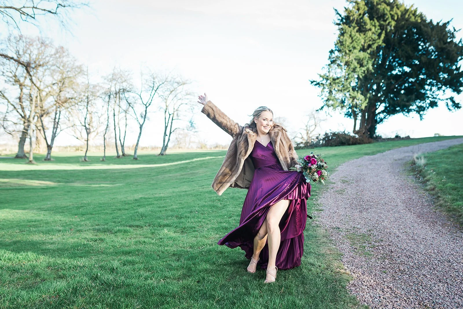 A woman in a purple dress and high heels, holding a bouquet of flowers, joyfully running on a grassy field with a dirt path, trees, and a clear sky in the background.