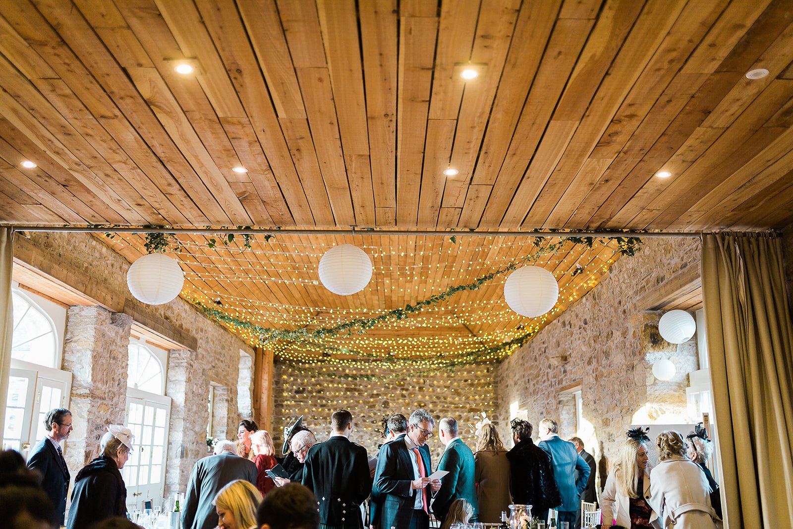 People gathered in a decorated rustic event hall with string lights, paper lanterns, and greenery on a wooden ceiling for a social gathering or celebration.