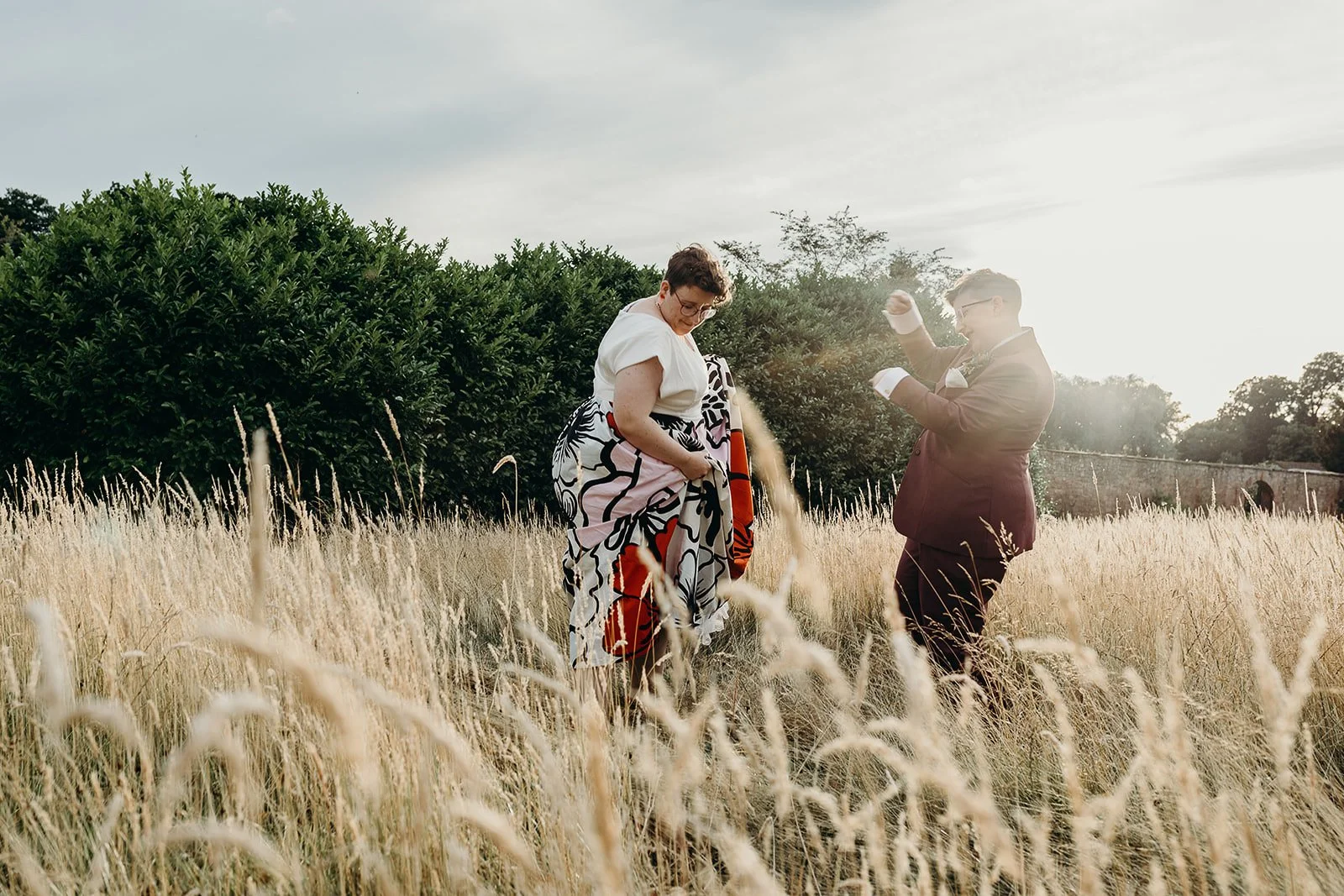 A couple in formal attire standing in a field of tall, dry grass during sunset, with the woman looking down and adjusting her dress, and the man looking at his phone, smiling.