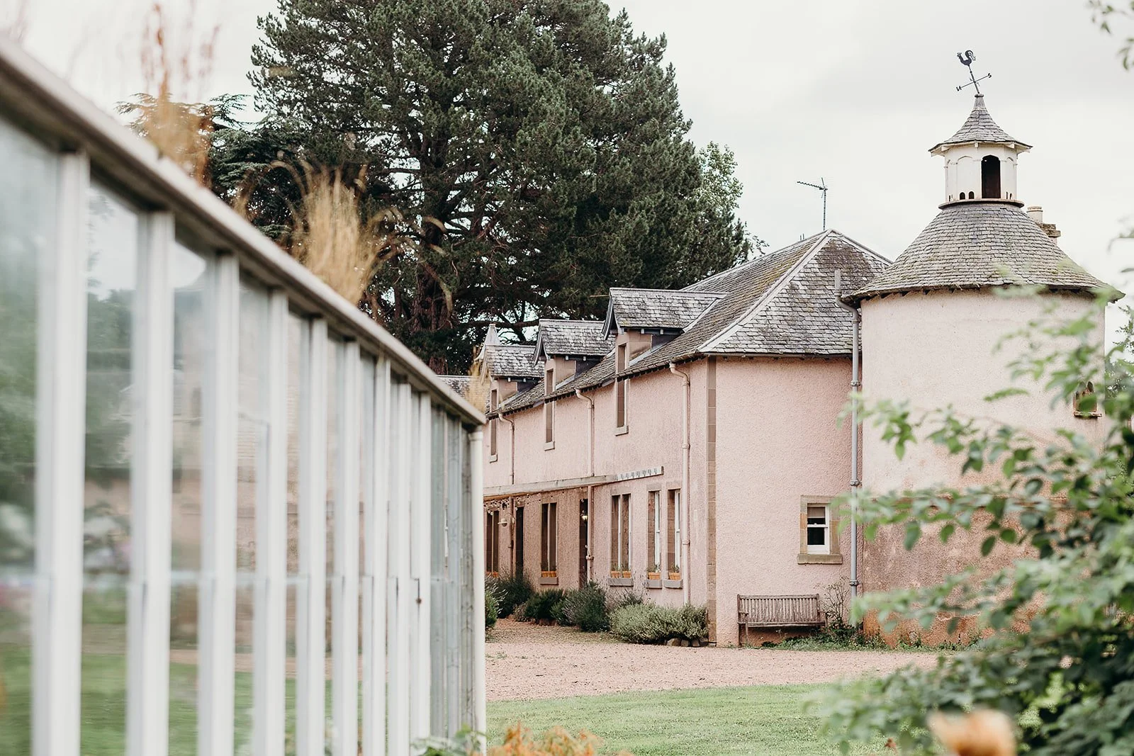 A pink building with a turret and multiple windows, surrounded by green grass and trees, viewed through a white fence.