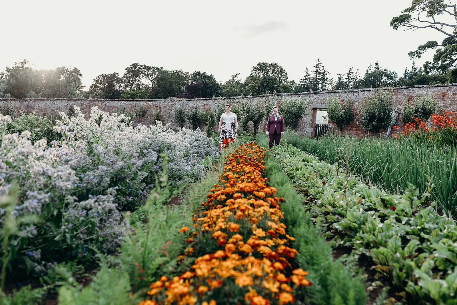 Two women walking through a lush garden with rows of colorful flowers and vegetables, surrounded by a brick wall and green trees in the background.