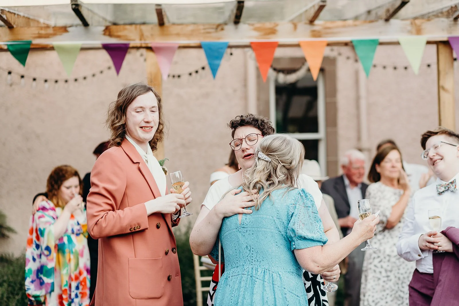 People celebrating at an outdoor gathering, some holding champagne glasses, with colorful bunting hanging overhead.