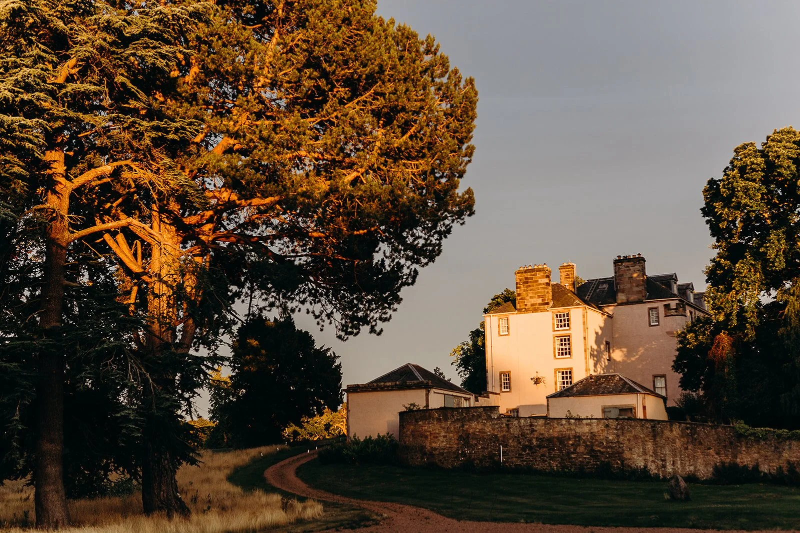 A historical house with multiple chimneys, surrounded by trees and a stone wall, with a dirt pathway leading up to it, during sunset.