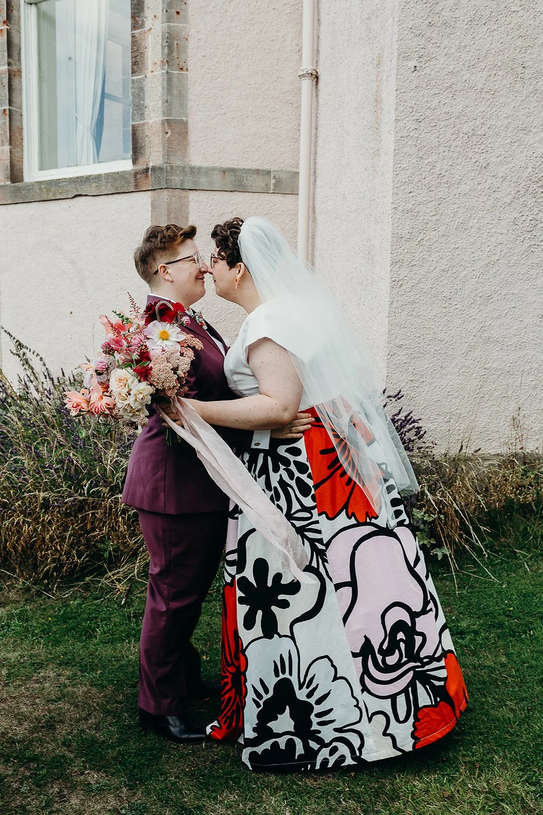 A same-sex couple in wedding attire sharing a intimate moment outdoors, one holding a bouquet of flowers, near a building with a window and garden plants.