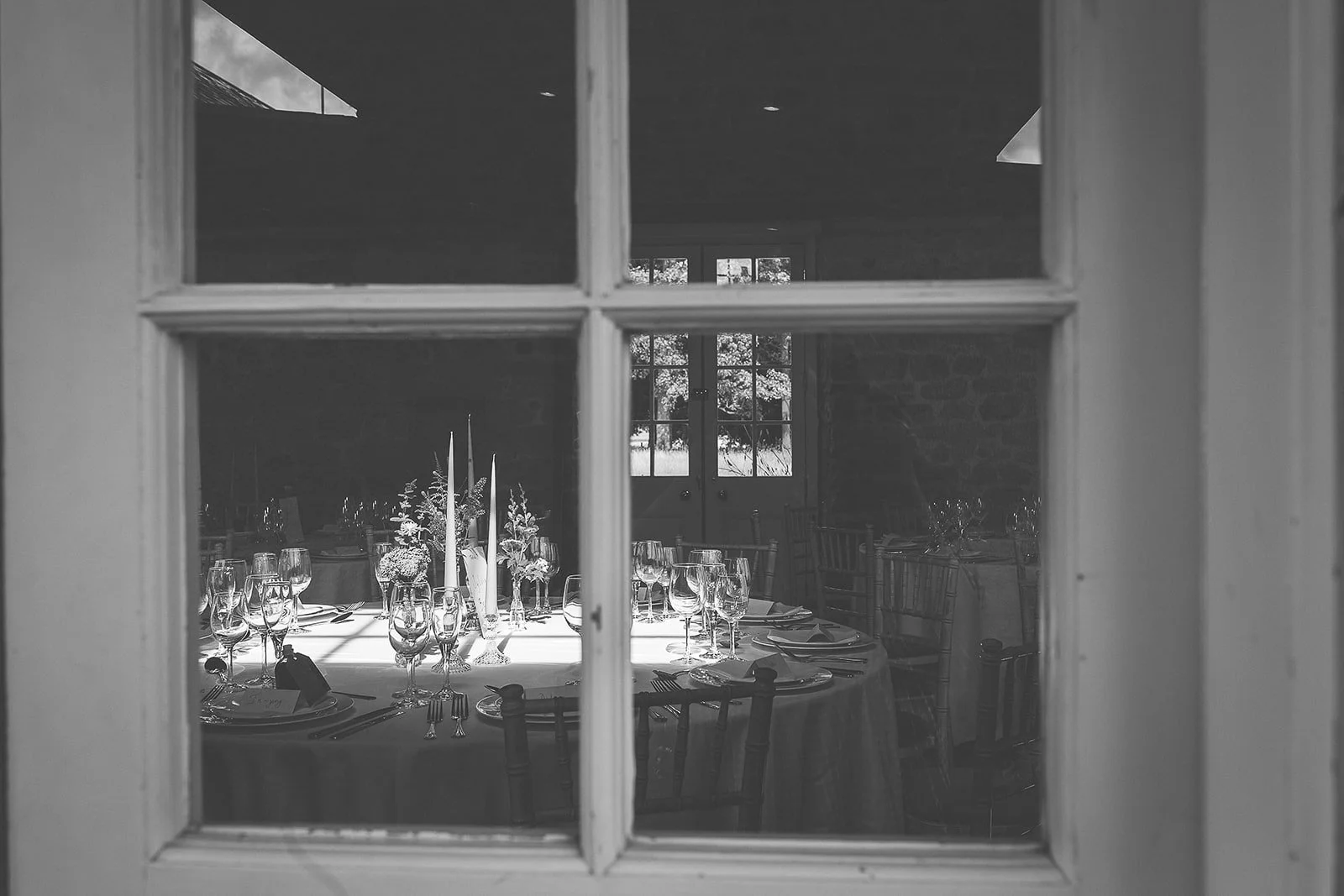 A dining room set up for a formal event, viewed through a window. The table is decorated with candles and floral arrangements and is set with glassware, plates, and silverware. Sunlight streams through a window at the far end of the room, illuminatin