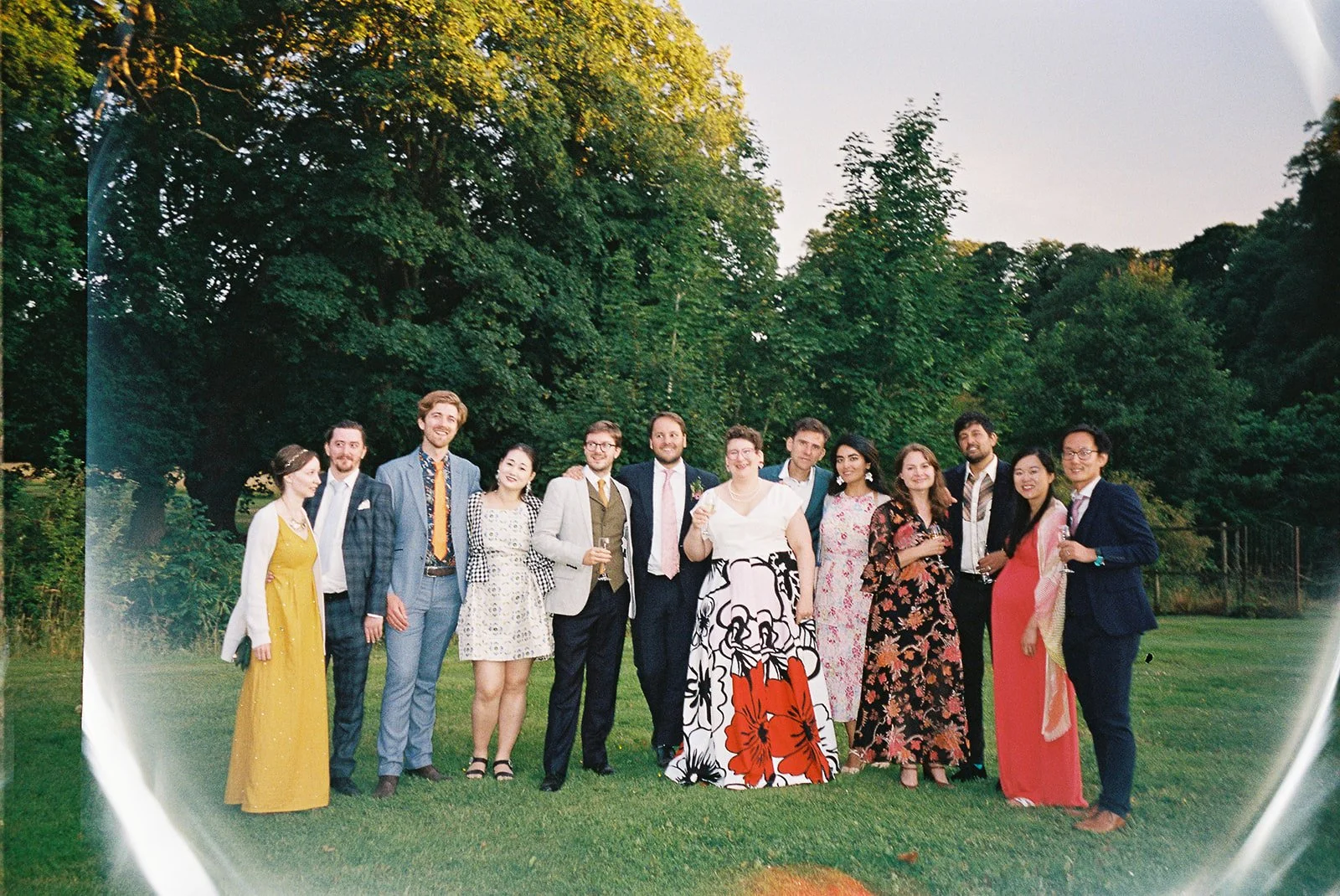 A group of 13 people dressed in formal attire posing outdoors on a grassy area with trees in the background during sunset.