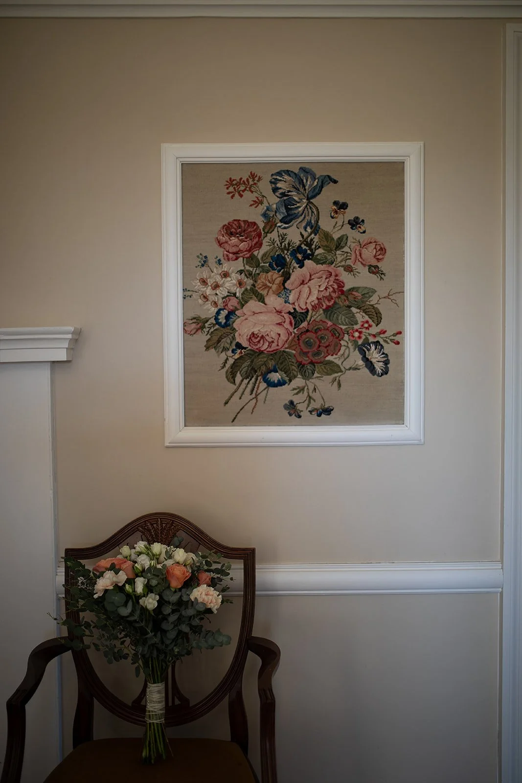A floral tapestry hangs on a beige wall above a wooden chair with a bouquet of flowers resting on it.