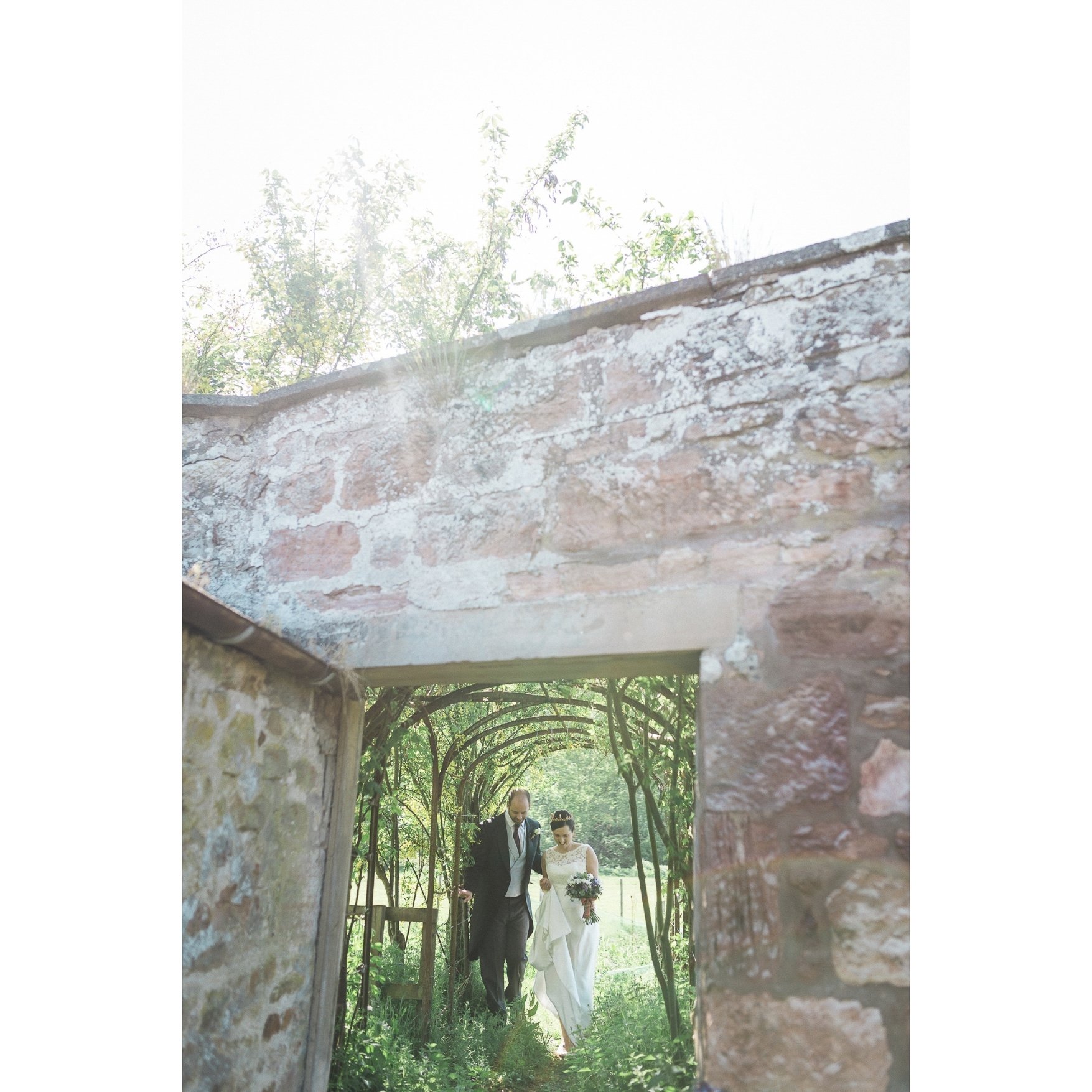 A bride and groom walking through a garden archway covered with greenery.