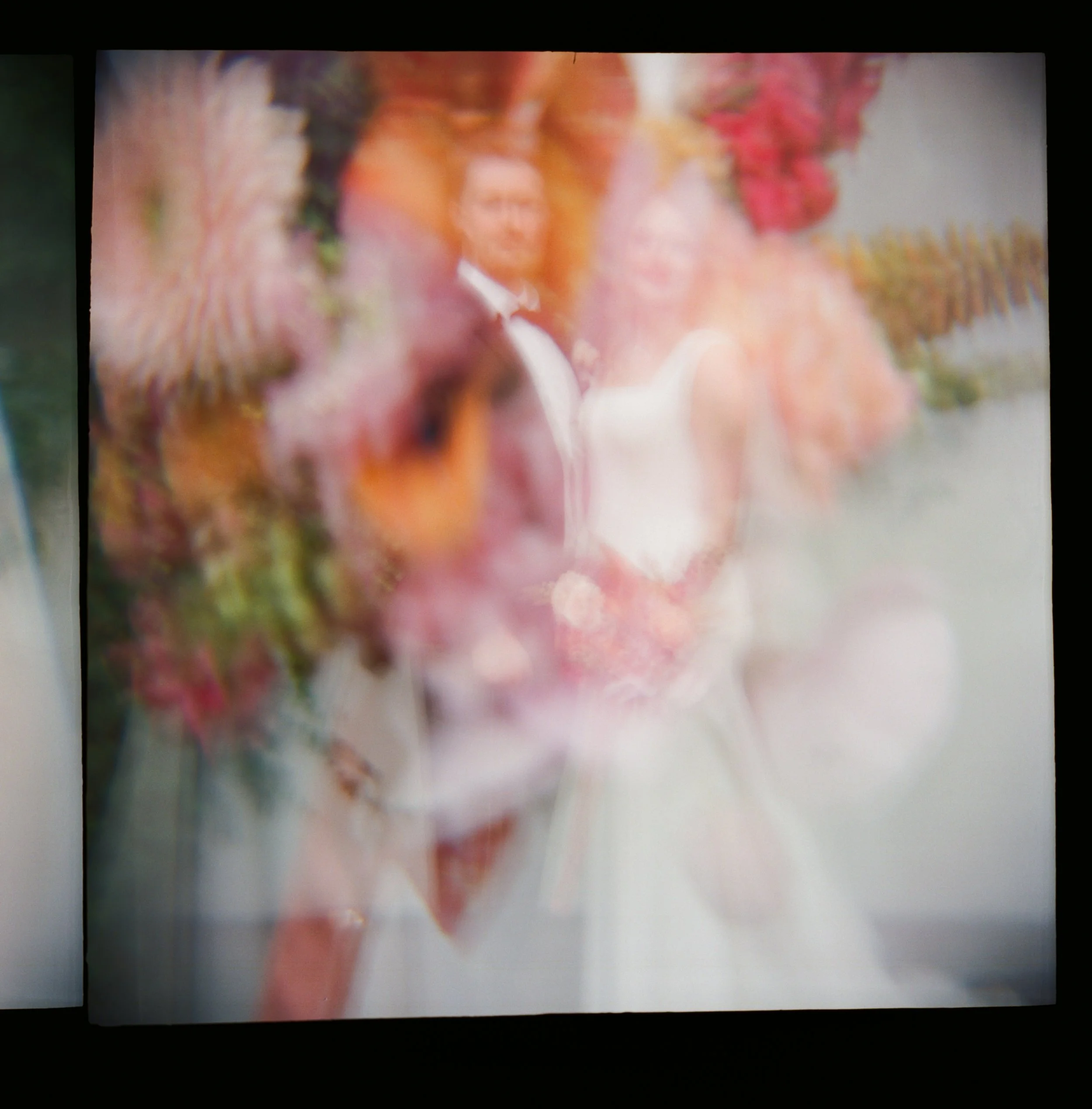Blurred photograph of a bride and groom in wedding attire surrounded by a floral display.