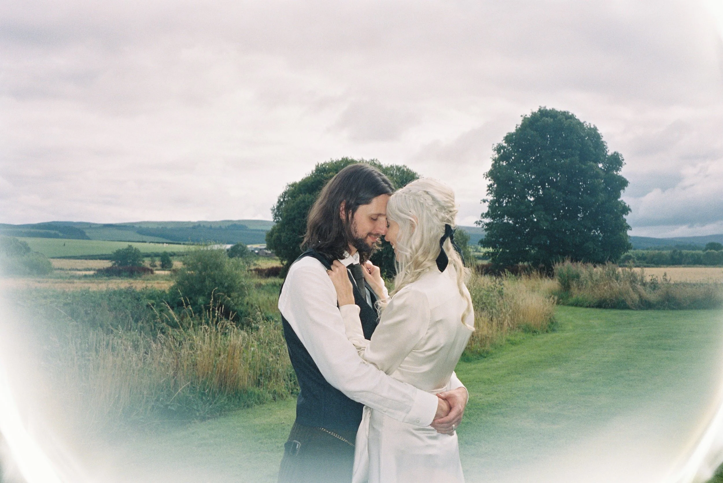A couple embracing outdoors on a cloudy day with green fields and trees in the background.