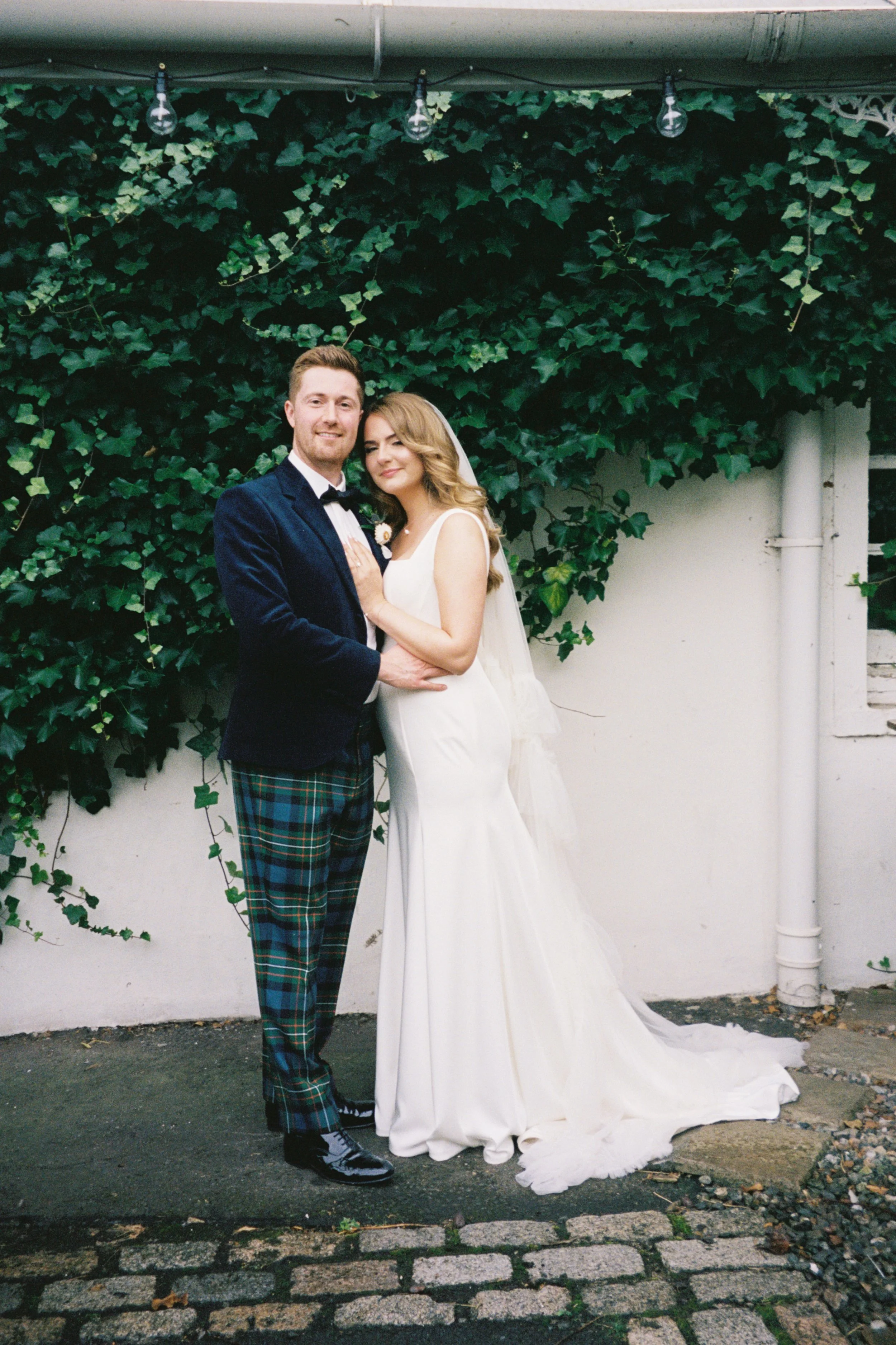 A newlywed couple standing outdoors in front of green foliage, with the groom in a dark blazer and plaid pants, and the bride in a white wedding gown, both smiling.