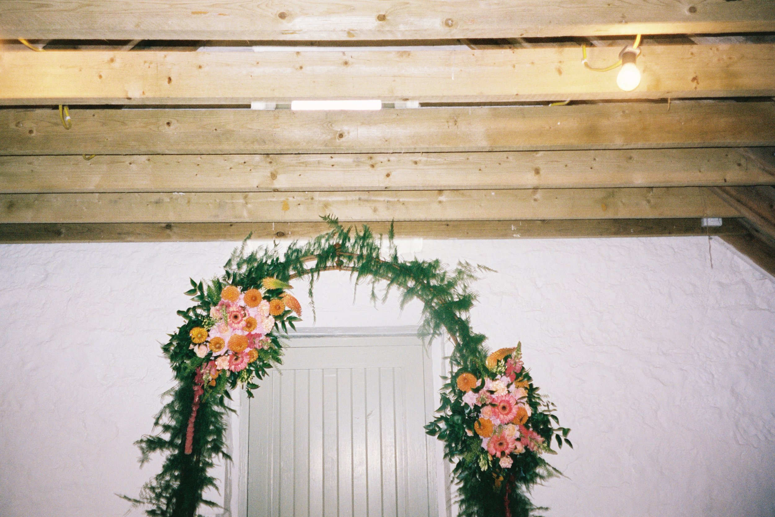 A floral arch with pink and orange flowers and green foliage, positioned over a closed door in an interior space with a white textured wall and wooden ceiling beams.