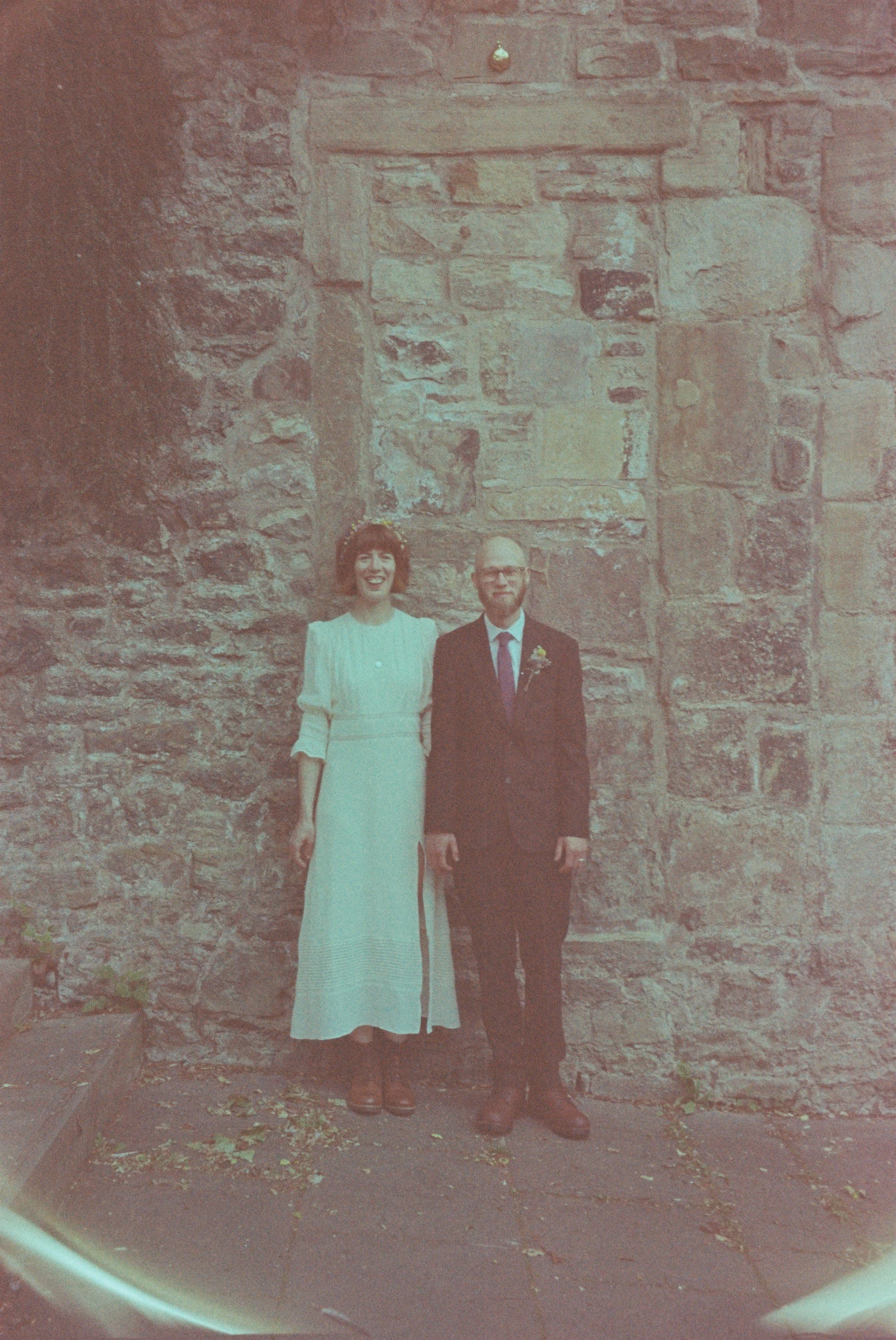 A woman in a white dress and a man in a black suit with a boutonniere, standing in front of a stone wall, smiling for a photo.