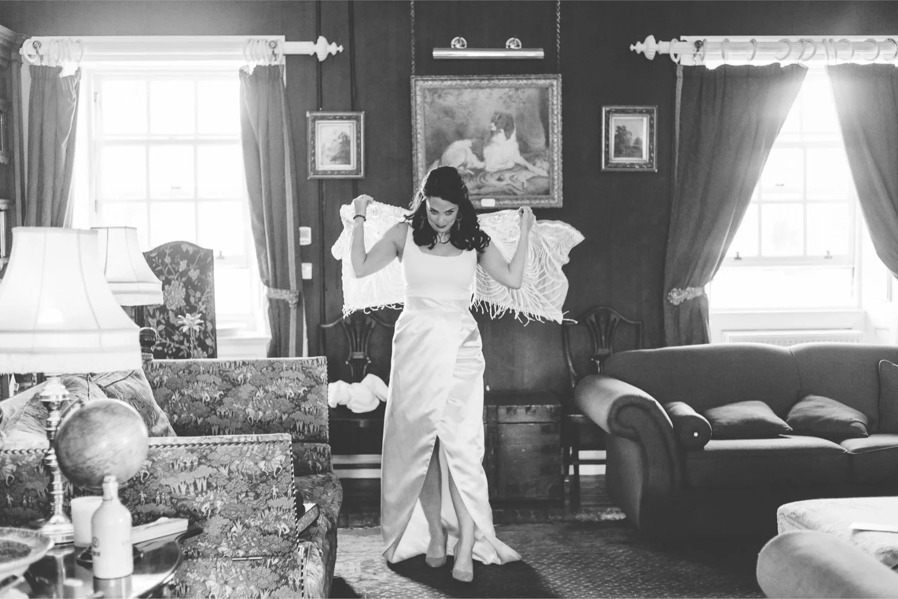 A woman in a white dress standing in a vintage living room, adjusting a shawl on her shoulders, with furniture and artwork around her.