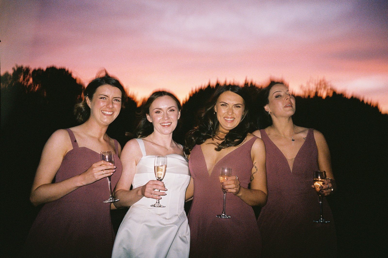 Four women in elegant dresses holding glasses of champagne at sunset outdoor gathering.