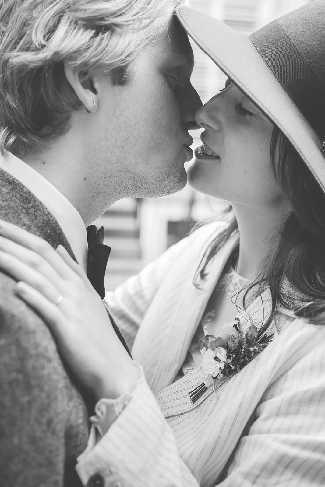 A close-up black-and-white photo of a couple about to kiss, dressed in formal attire, with the woman wearing a hat and a small flower boutonniere.