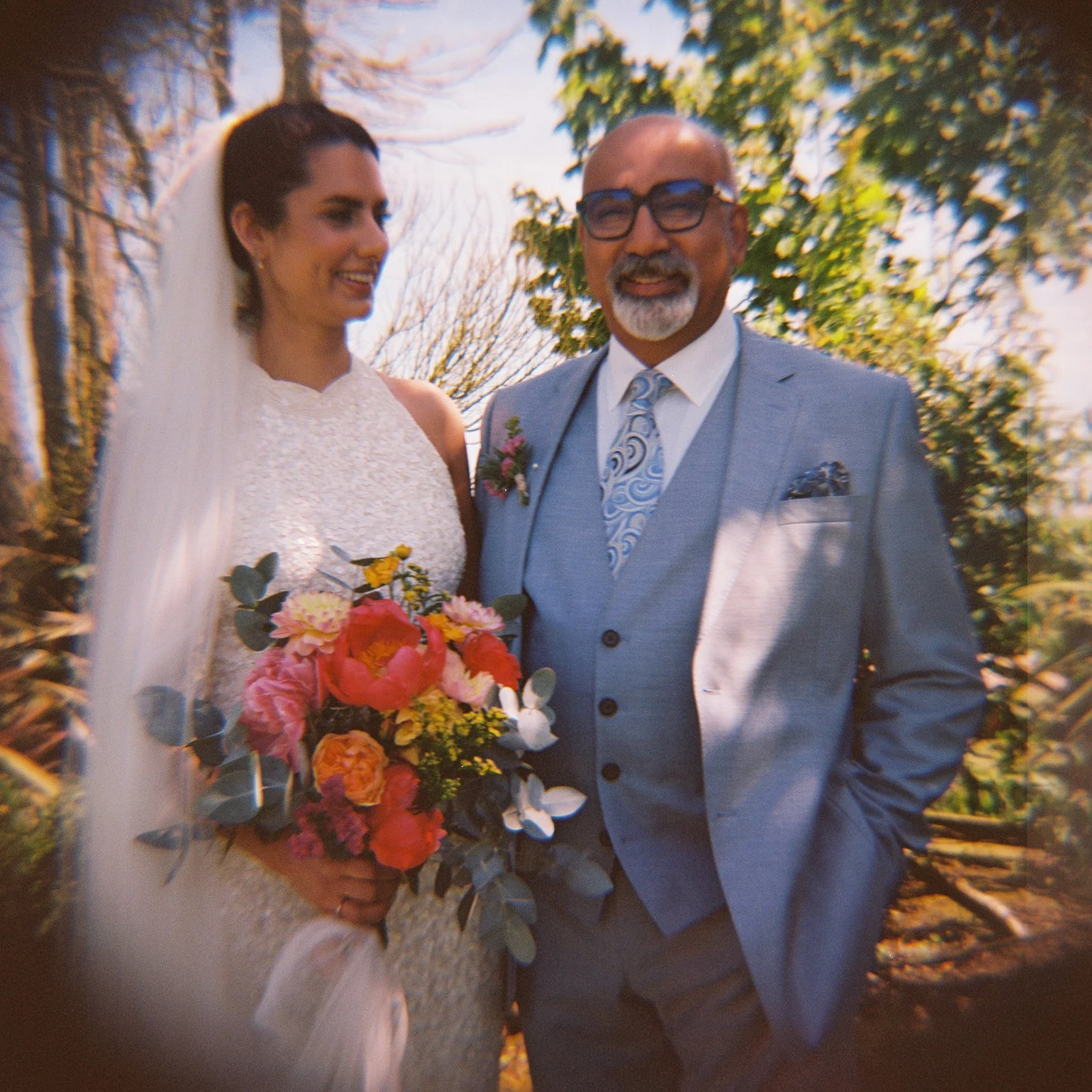 A bride and a man in a suit smiling outdoors on a sunny day. The bride holds a colorful bouquet of flowers, and they are surrounded by trees with budding leaves.