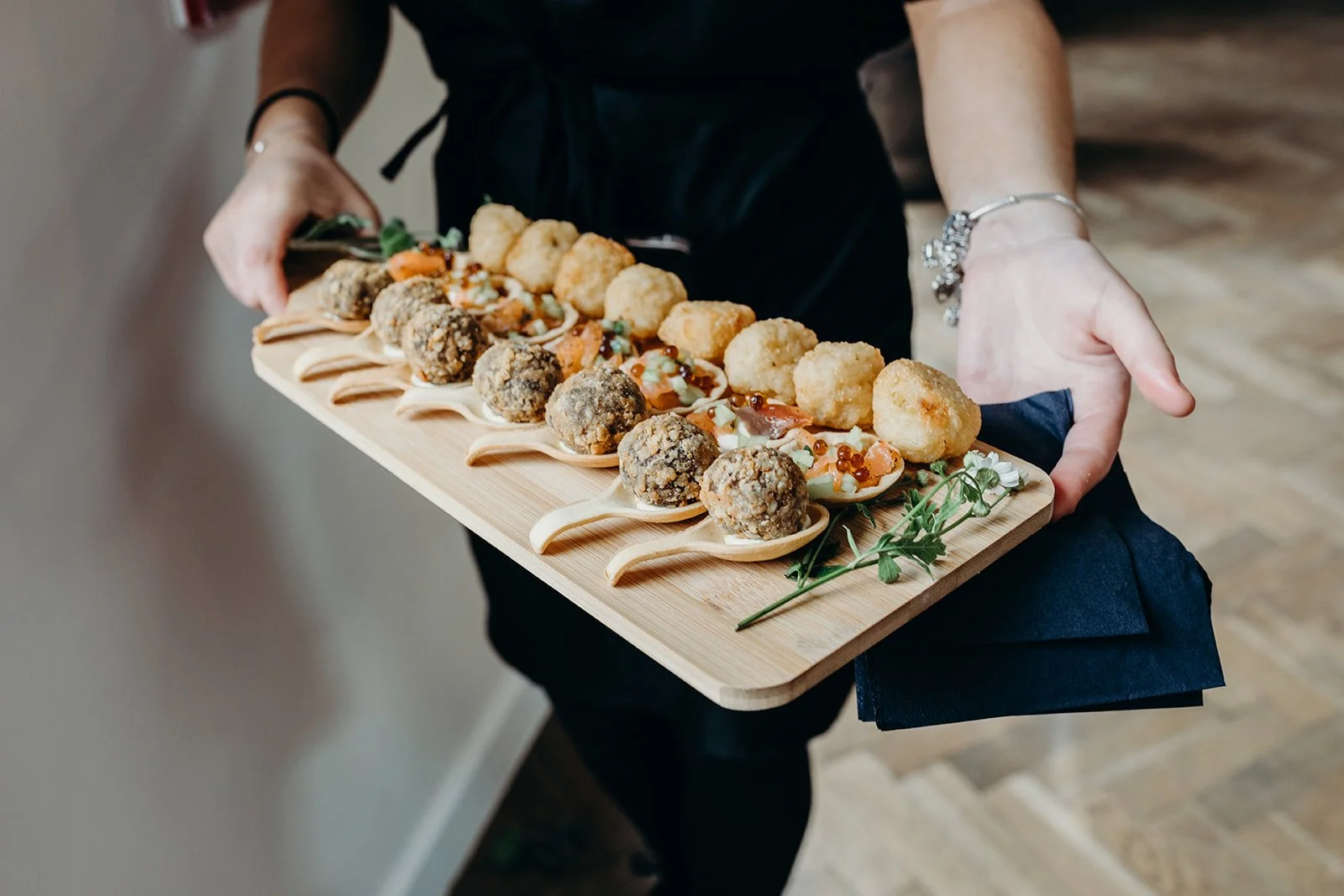 A person holding a wooden tray with assorted appetizer bites, including meatballs, croquettes, and small edible bowls topped with sauce and garnishes.