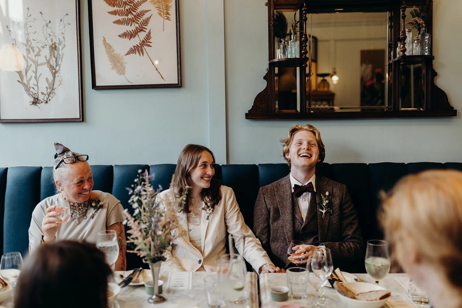People sitting at a dining table with drinks and flowers, smiling and laughing during a celebration or gathering.
