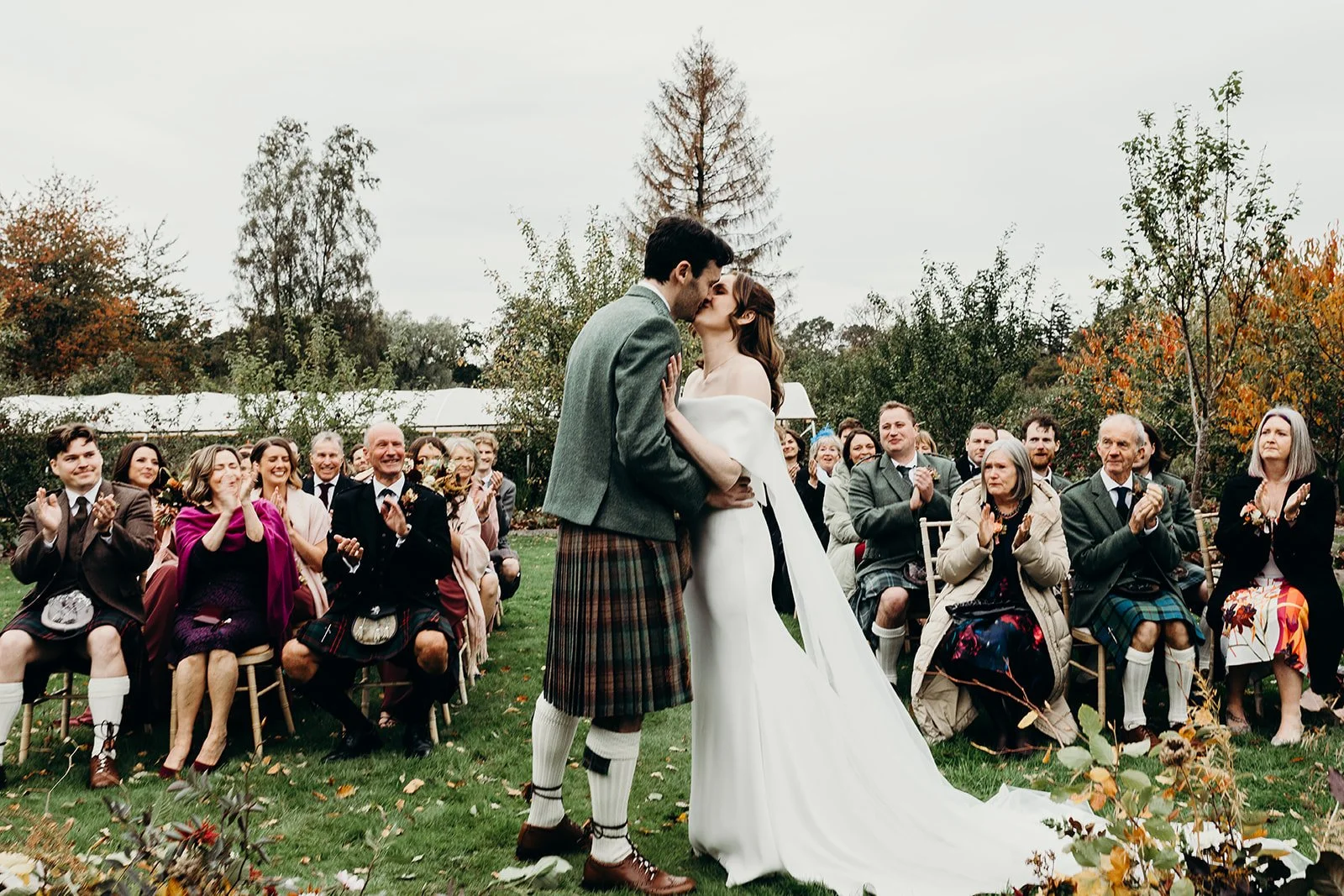 A wedding ceremony outdoors with a bride and groom kissing, surrounded by seated guests clapping and celebrating, with autumn trees in the background.