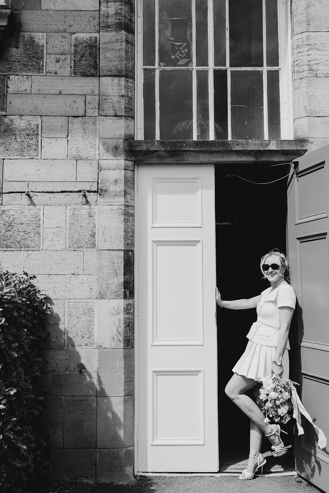 A woman in sunglasses, a light-colored dress, and high heels stands in a doorway holding a bouquet of flowers, smiling.