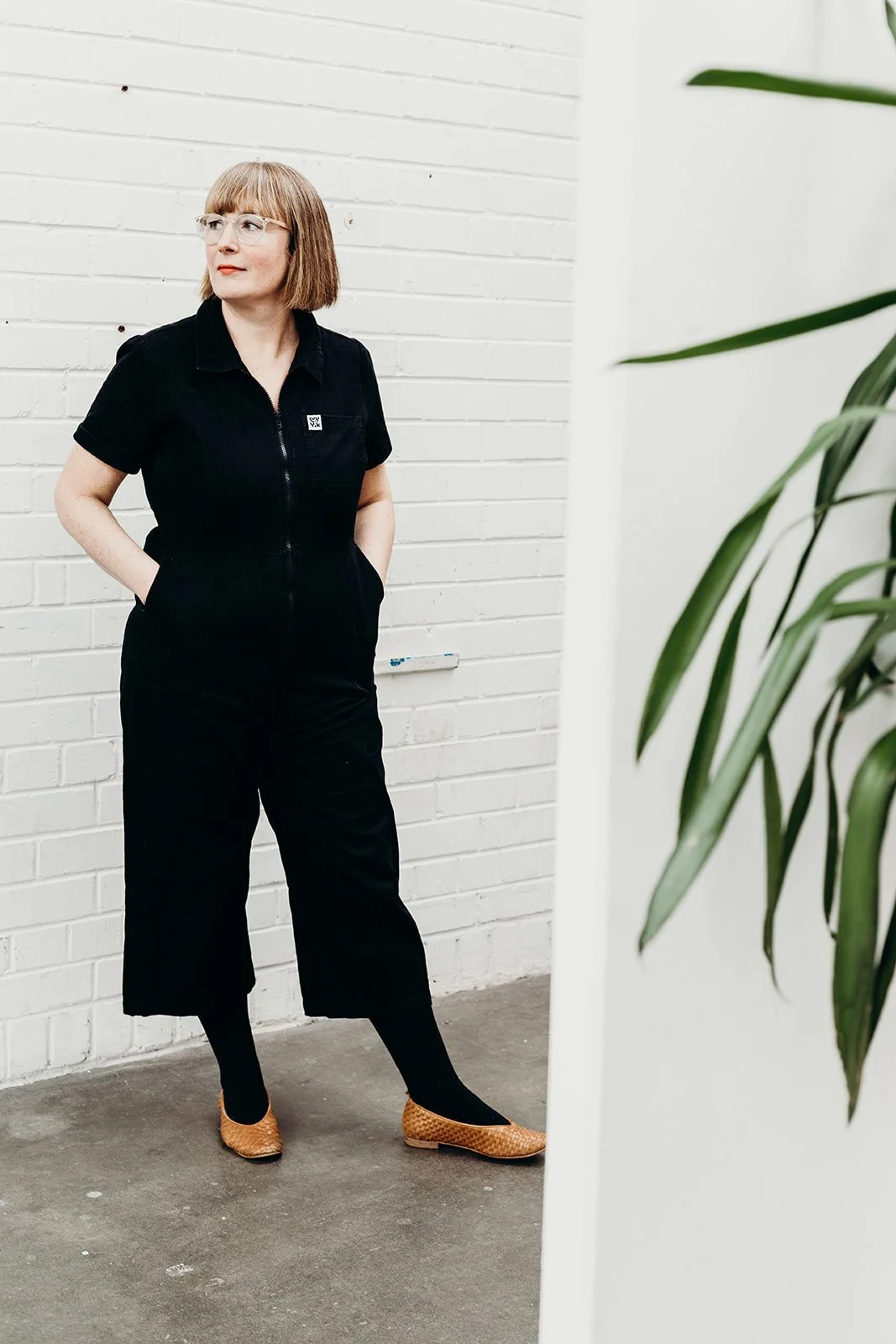 A woman with short light brown hair and glasses, wearing a black zip-up jumpsuit and tan woven shoes, standing against a white brick wall with her hands in her pockets, partially obscured by a white wall with green leaves in the foreground.