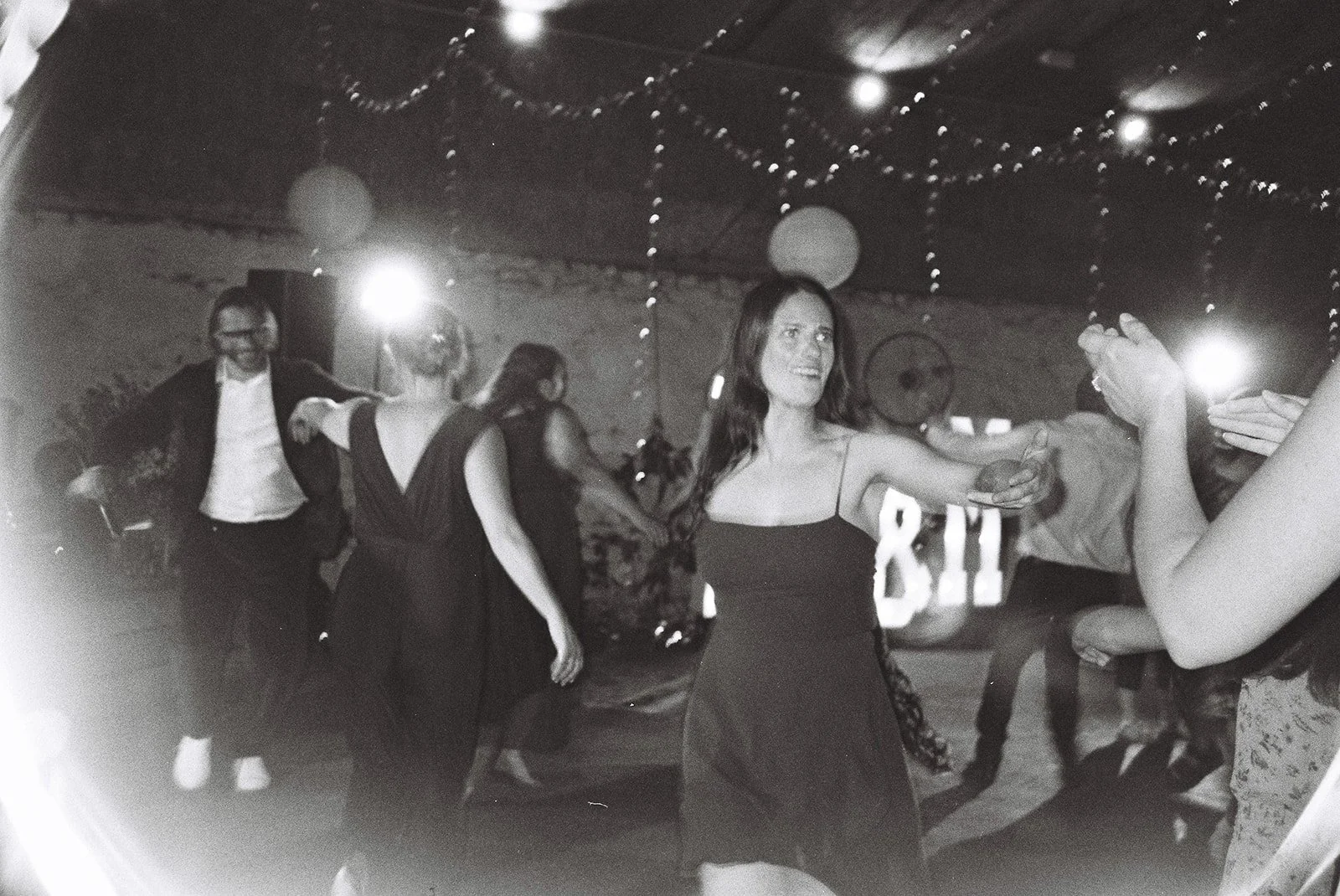 People dancing and socializing at a party with string lights and paper lanterns hanging from the ceiling.