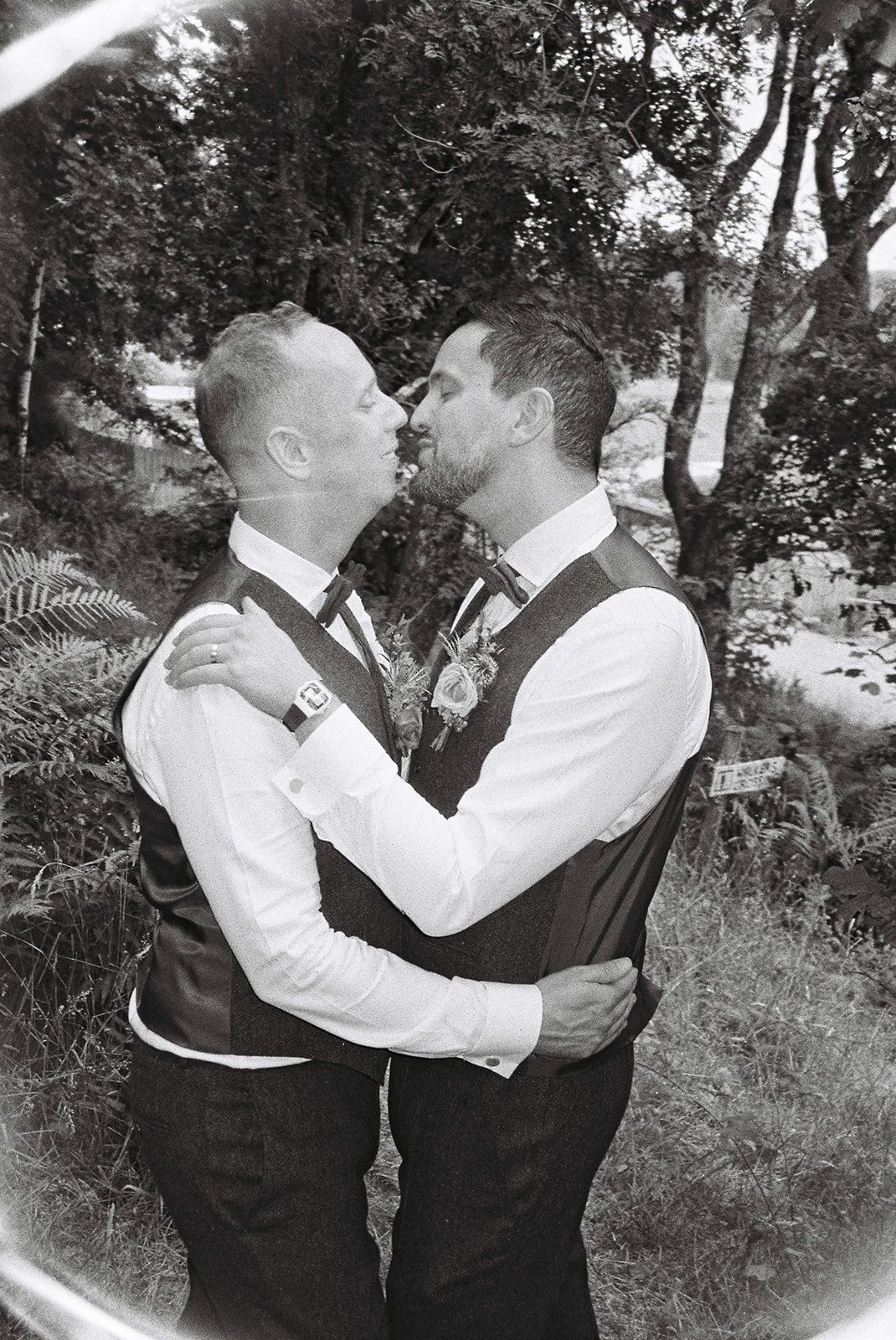 Two men dressed in formal vests and bow ties sharing a kiss outdoors, surrounded by trees and foliage in a black-and-white photograph.