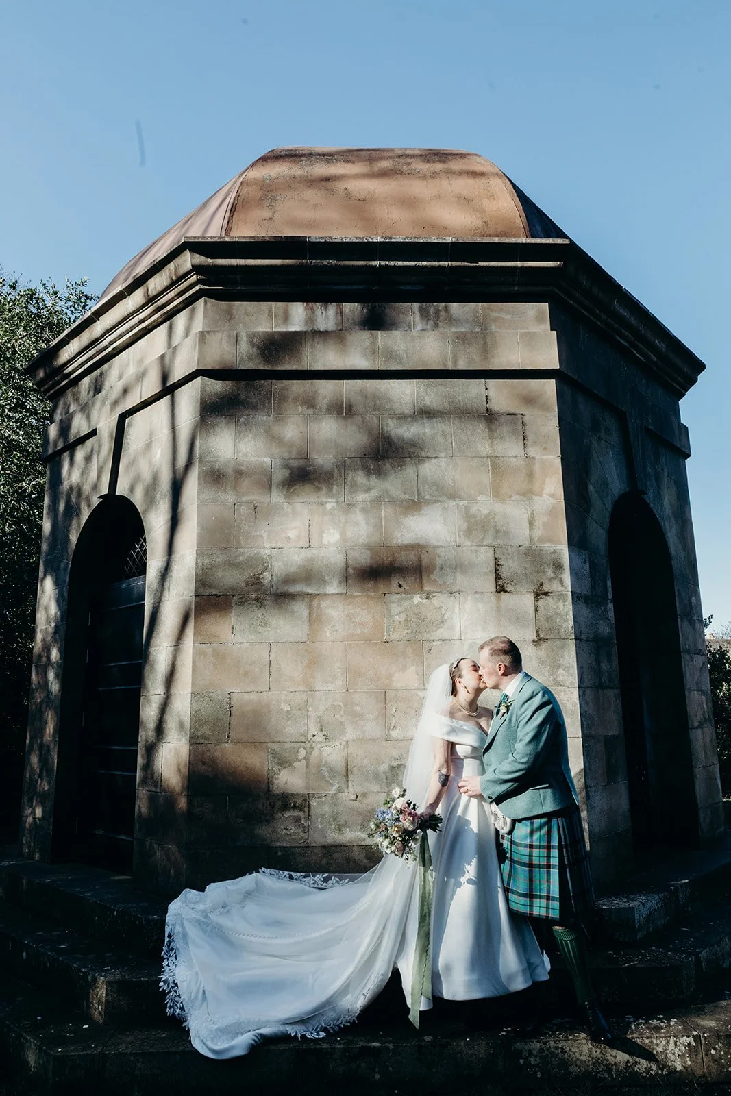 A bride and groom sharing a kiss in front of a stone monument on a clear day