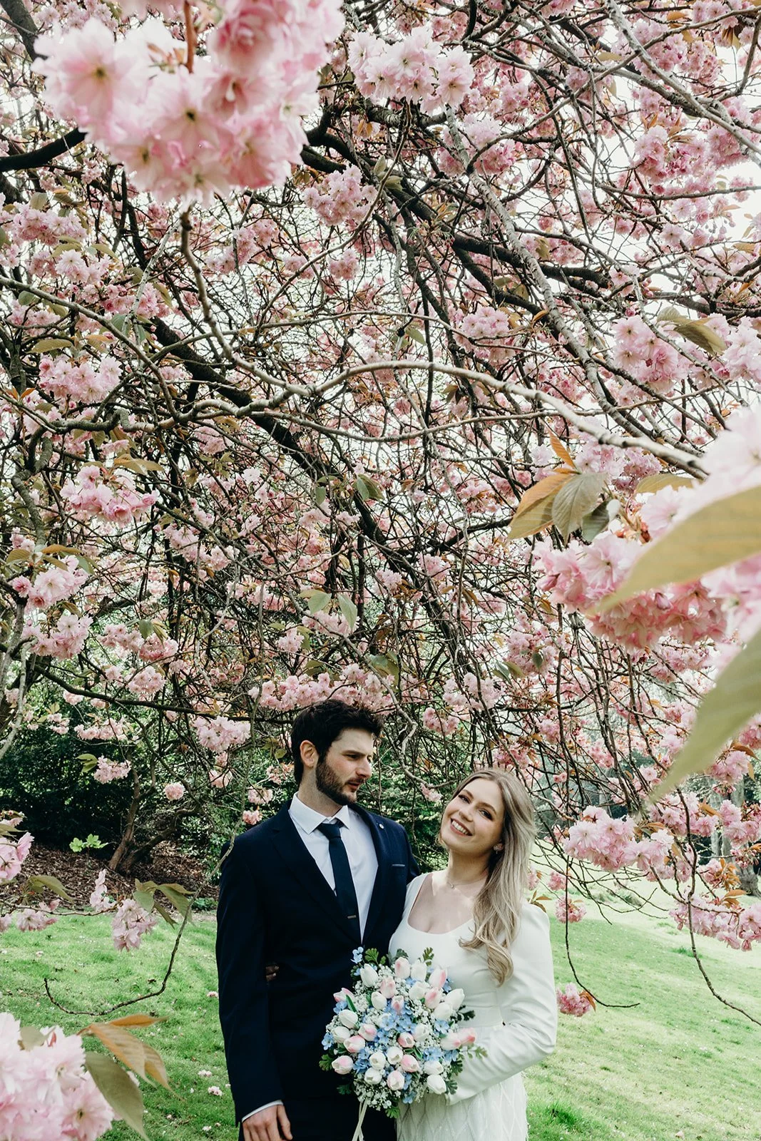 A couple in wedding attire stands under pink flowering cherry blossom trees, with the bride holding a bouquet and smiling.