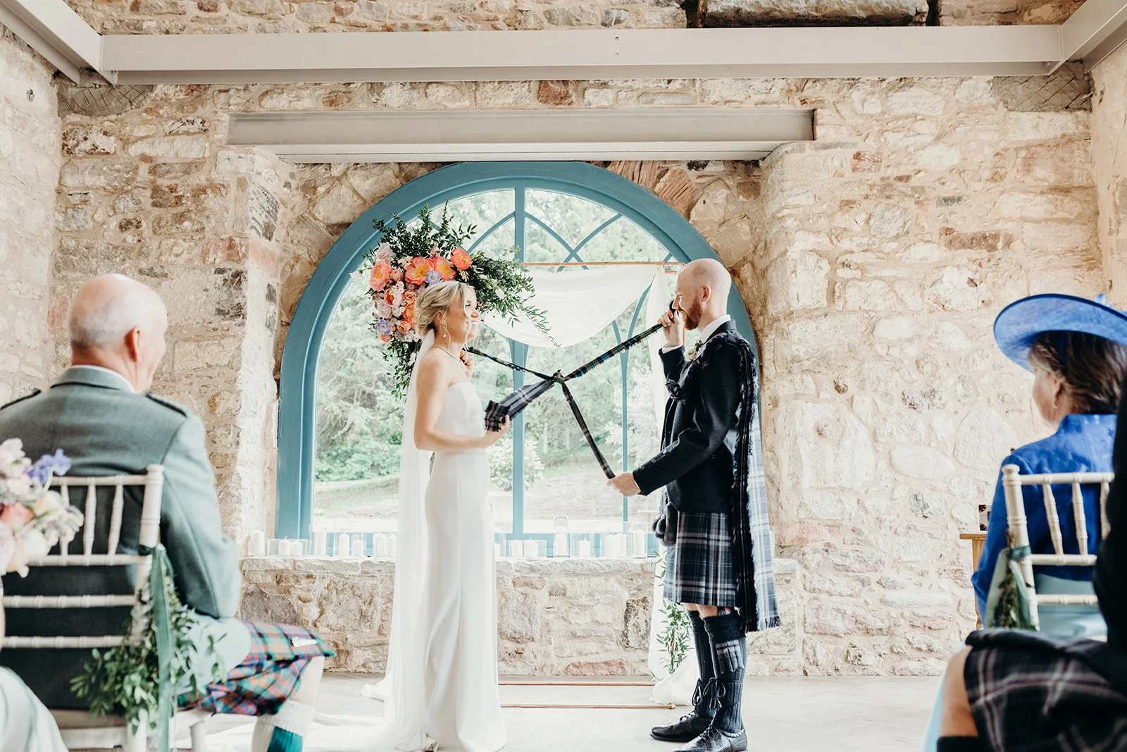 A wedding ceremony taking place in a rustic stone venue with a large blue arched window in the background. The bride and groom, both holding traditional Scottish bagpipes, are exchanging vows. Flower arrangements and guests in formal attire, including someone in a blue hat, are visible.