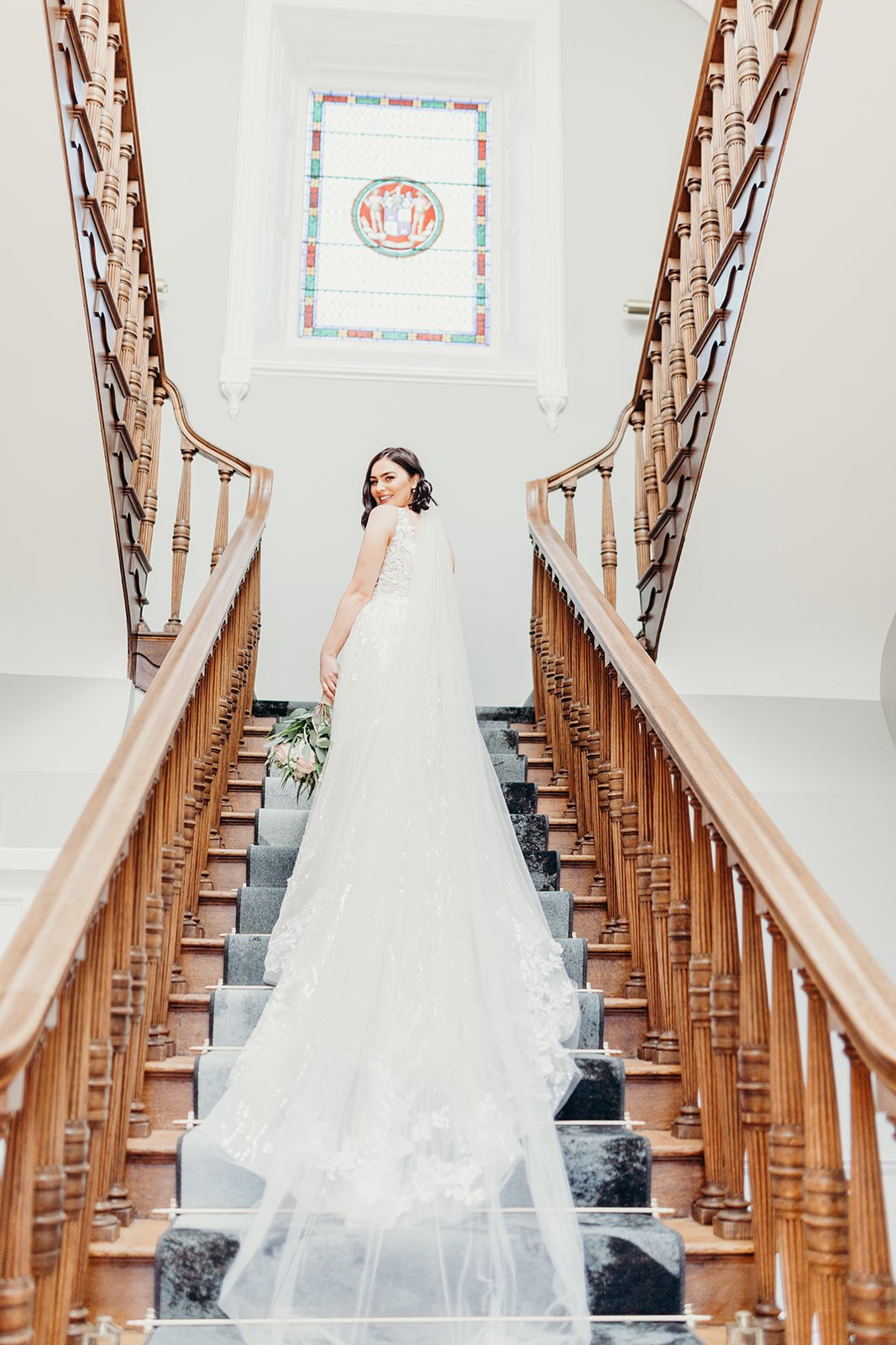 Bride in a white wedding gown standing on a staircase holding a bouquet, smiling at the camera, with a stained glass window overhead.
