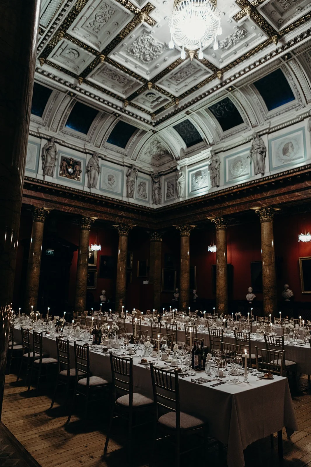 A grand, ornately decorated banquet hall with a high ceiling, chandelier, classical statues, red walls, marble columns, and a long formal dining table set with white tablecloths, candles, glassware, and silverware.