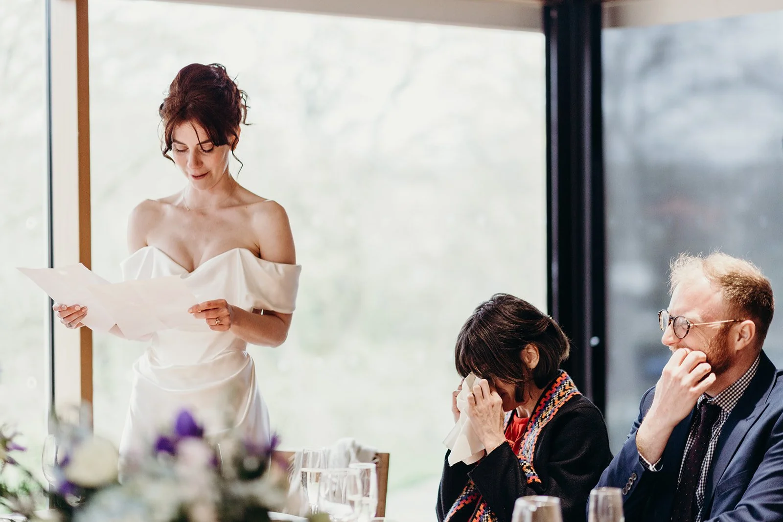 A woman in a white off-shoulder dress reading a paper at a table during a gathering, while two other people, a woman and a man, are seated and covering their faces, possibly crying, in a bright room with large windows.