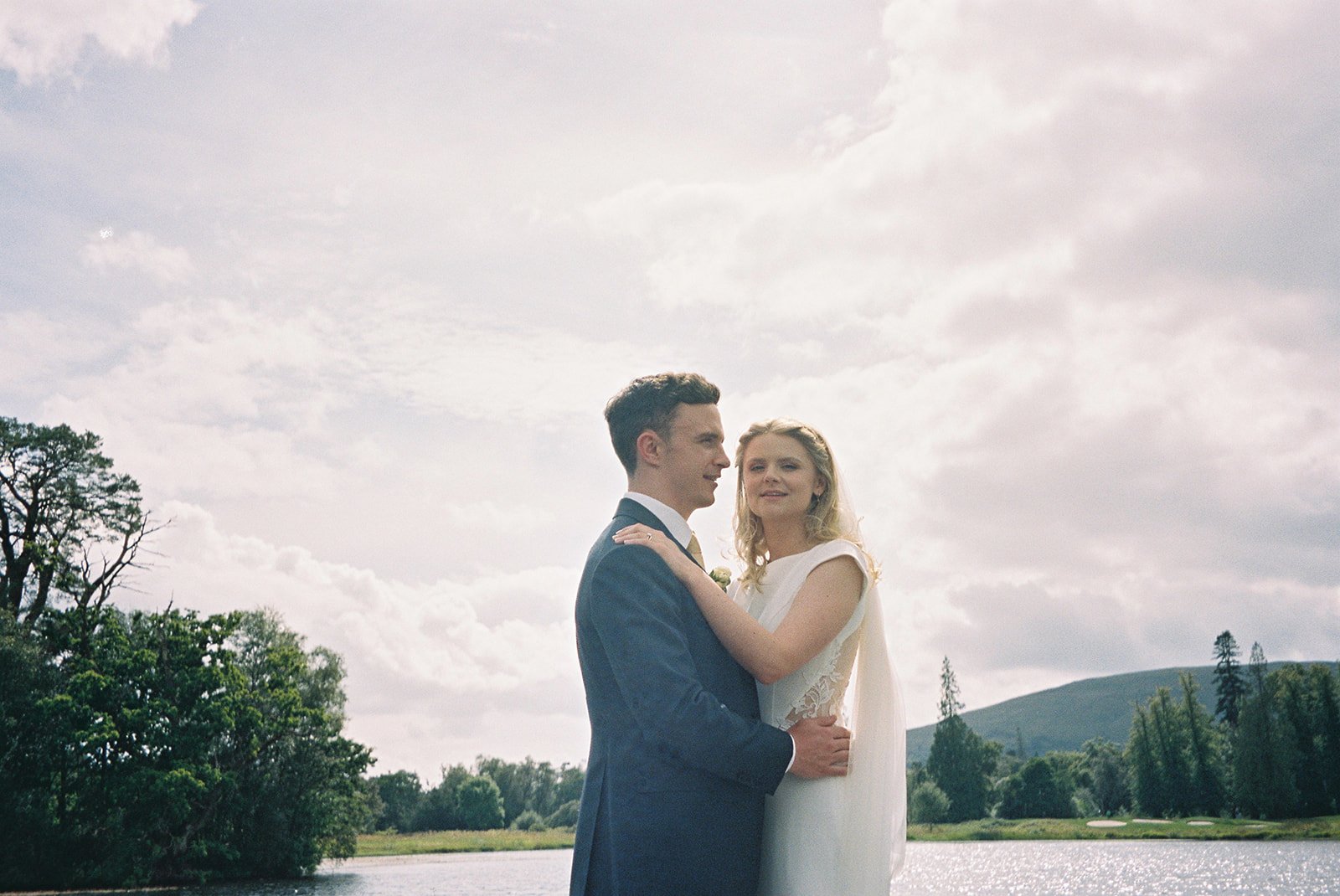 A newlywed couple standing by a lake with trees and mountains in the background, the man in a suit and the woman in a white dress, holding each other and smiling.